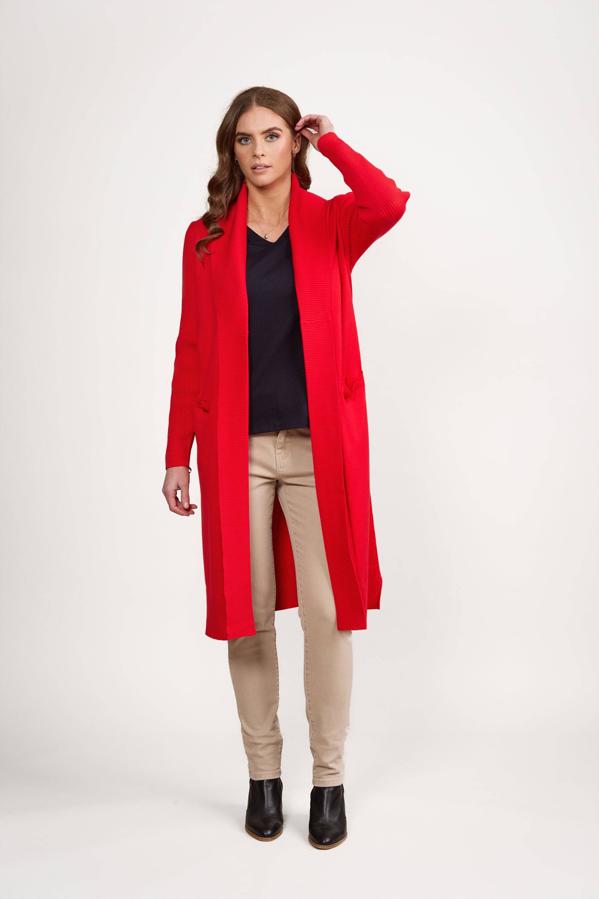 A woman stands against a white background wearing a long red cardigan, a navy top, beige pants, and black ankle boots. She has brown wavy hair and is touching her head with one hand.