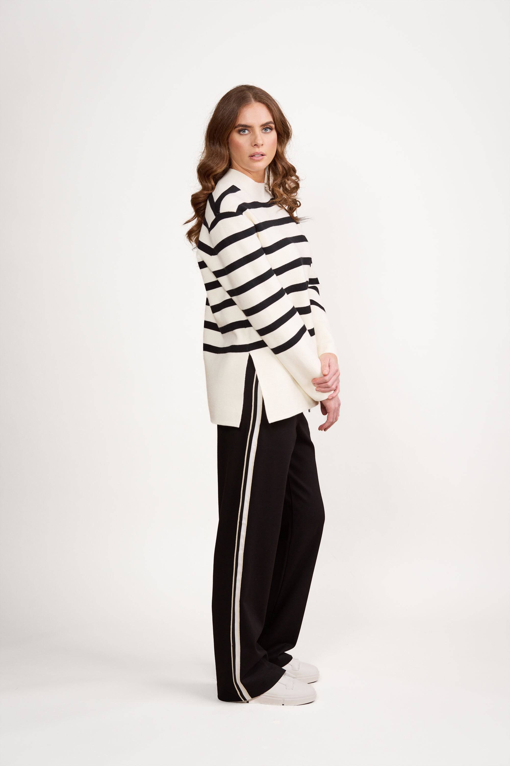Woman with wavy brown hair wearing a white and black striped sweater, black pants with a white side stripe, and white shoes, standing sideways against a plain white background.