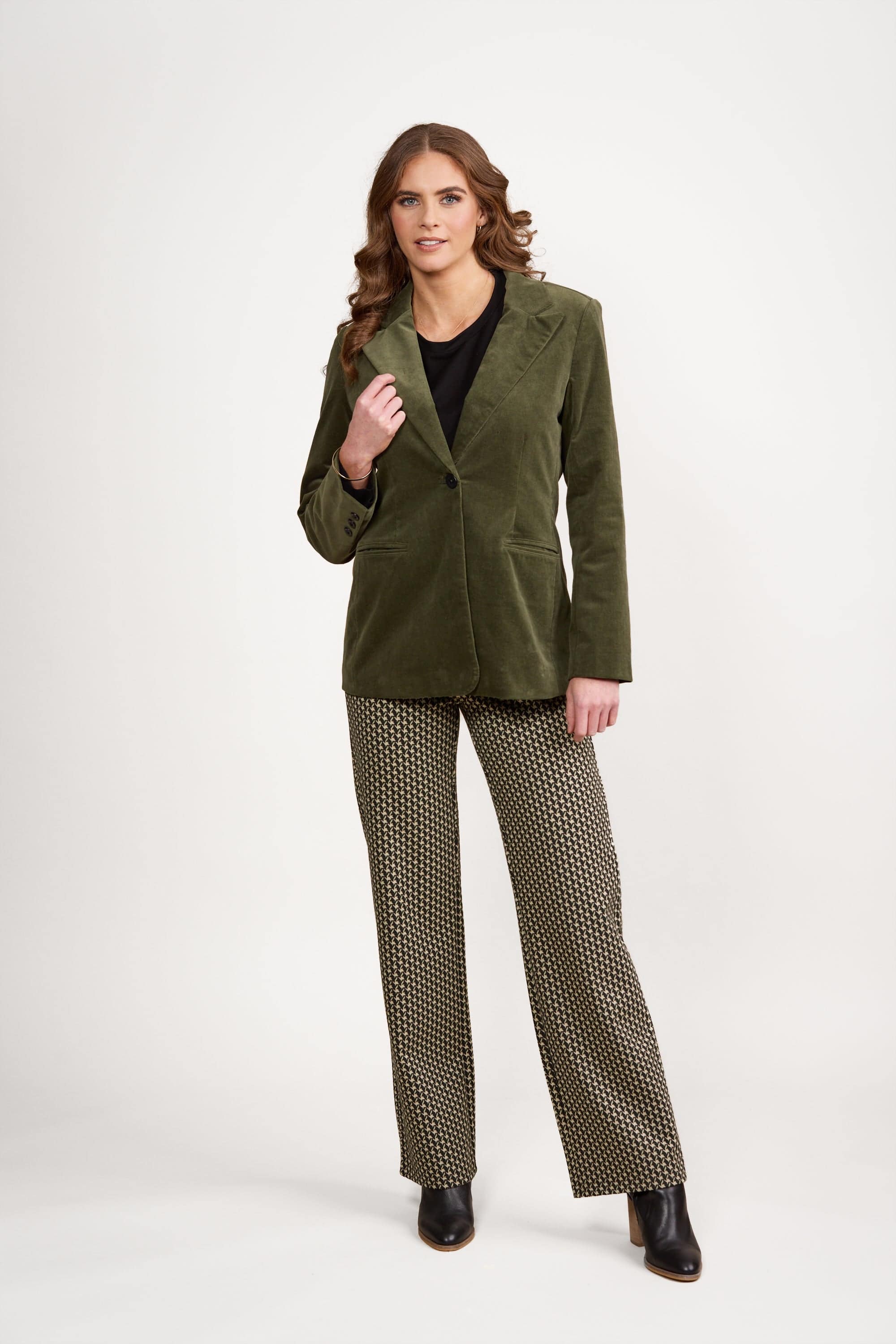 A woman stands against a white background wearing an olive green blazer, black shirt, patterned wide-leg trousers, and black ankle boots. She has wavy brown hair and a neutral expression.