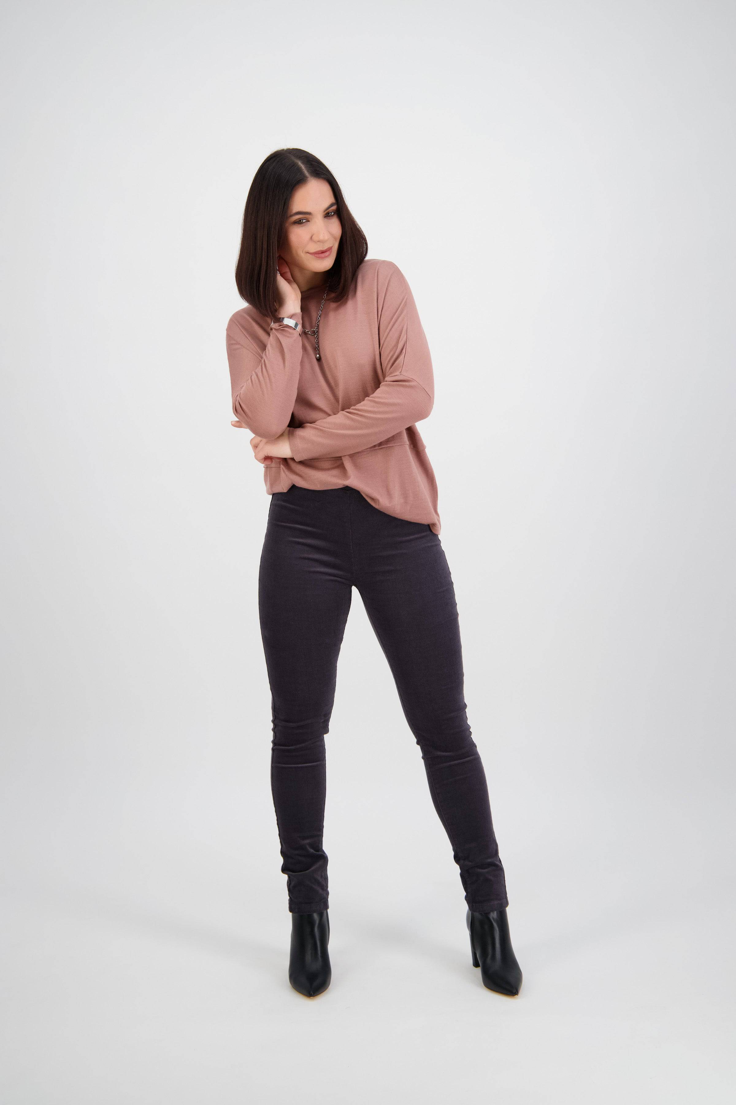 Woman with shoulder-length dark hair wearing a pink long-sleeve top, dark pants, and black boots stands with legs apart and arms crossed, smiling in front of a plain white background.