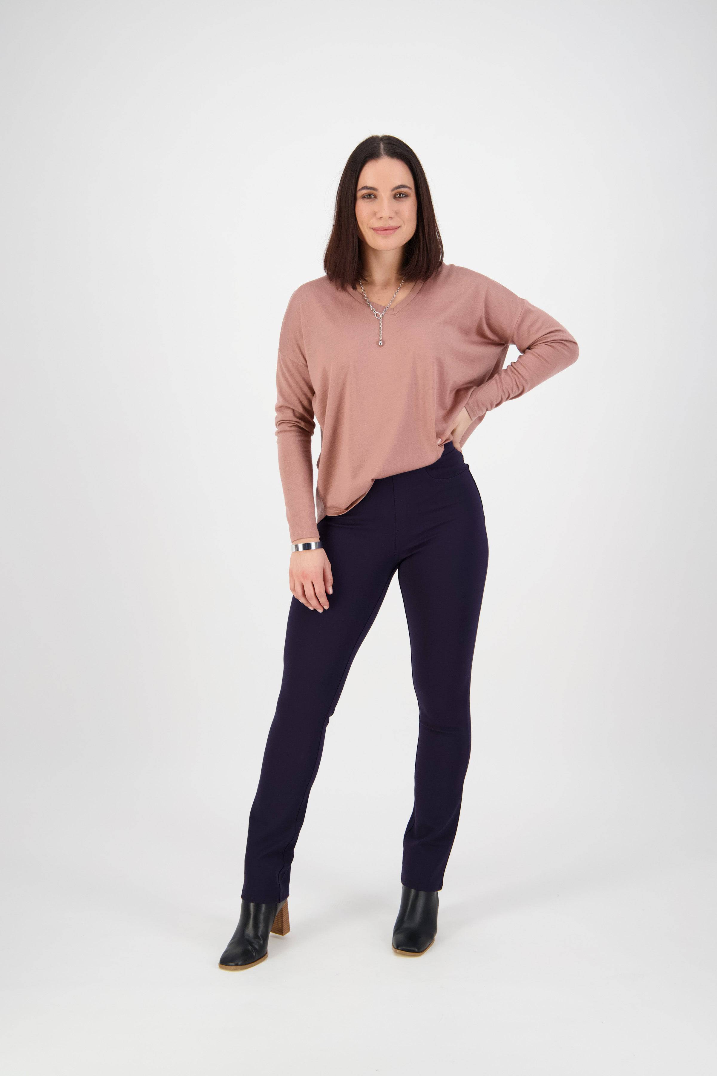 A woman with straight shoulder-length brown hair stands confidently, wearing a mauve long-sleeve top, navy pants, and black heeled boots against a plain white background.