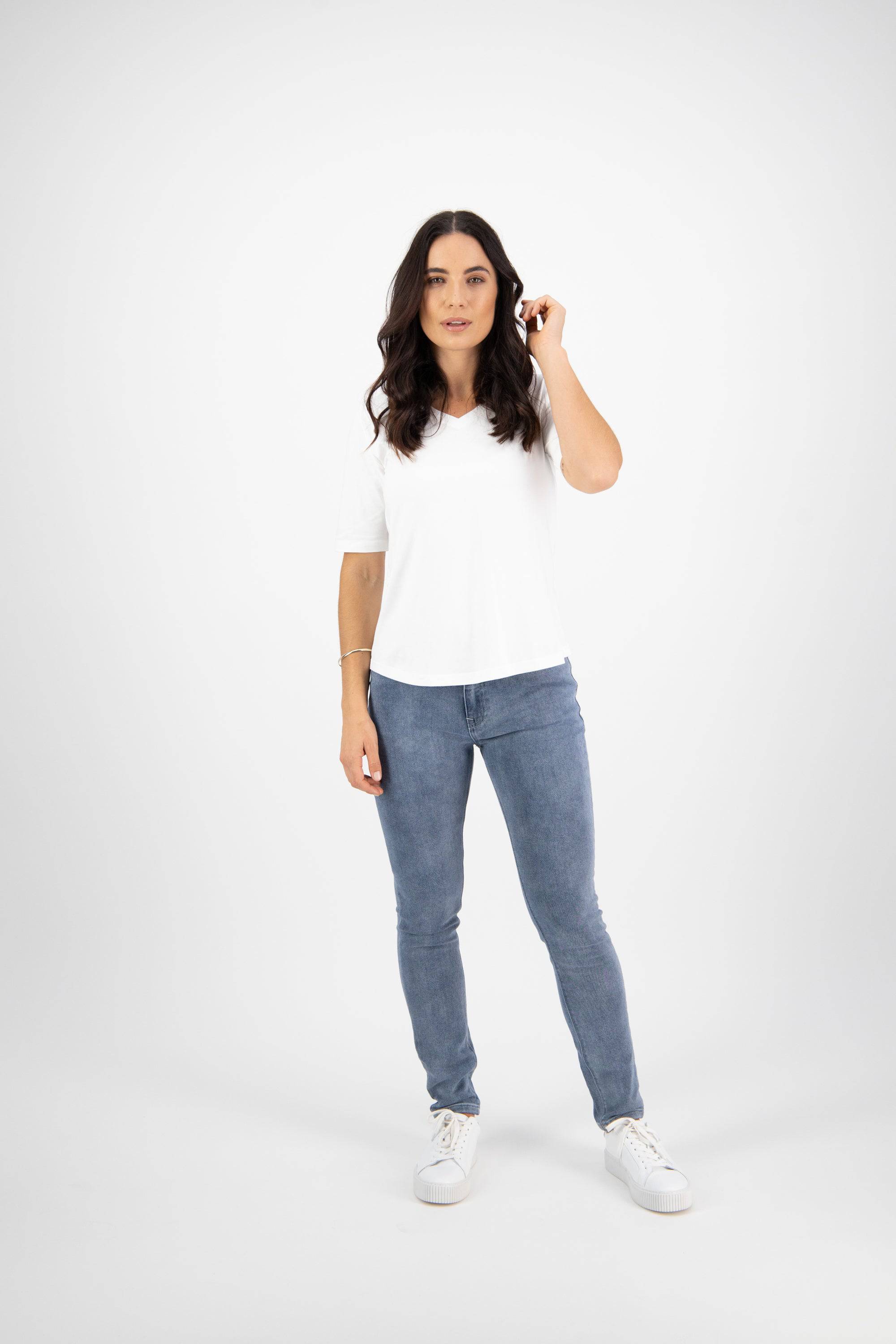 A woman with long dark hair stands against a white background, wearing a white t-shirt, blue jeans, and white sneakers. She has a relaxed expression and holds one hand near her hair.