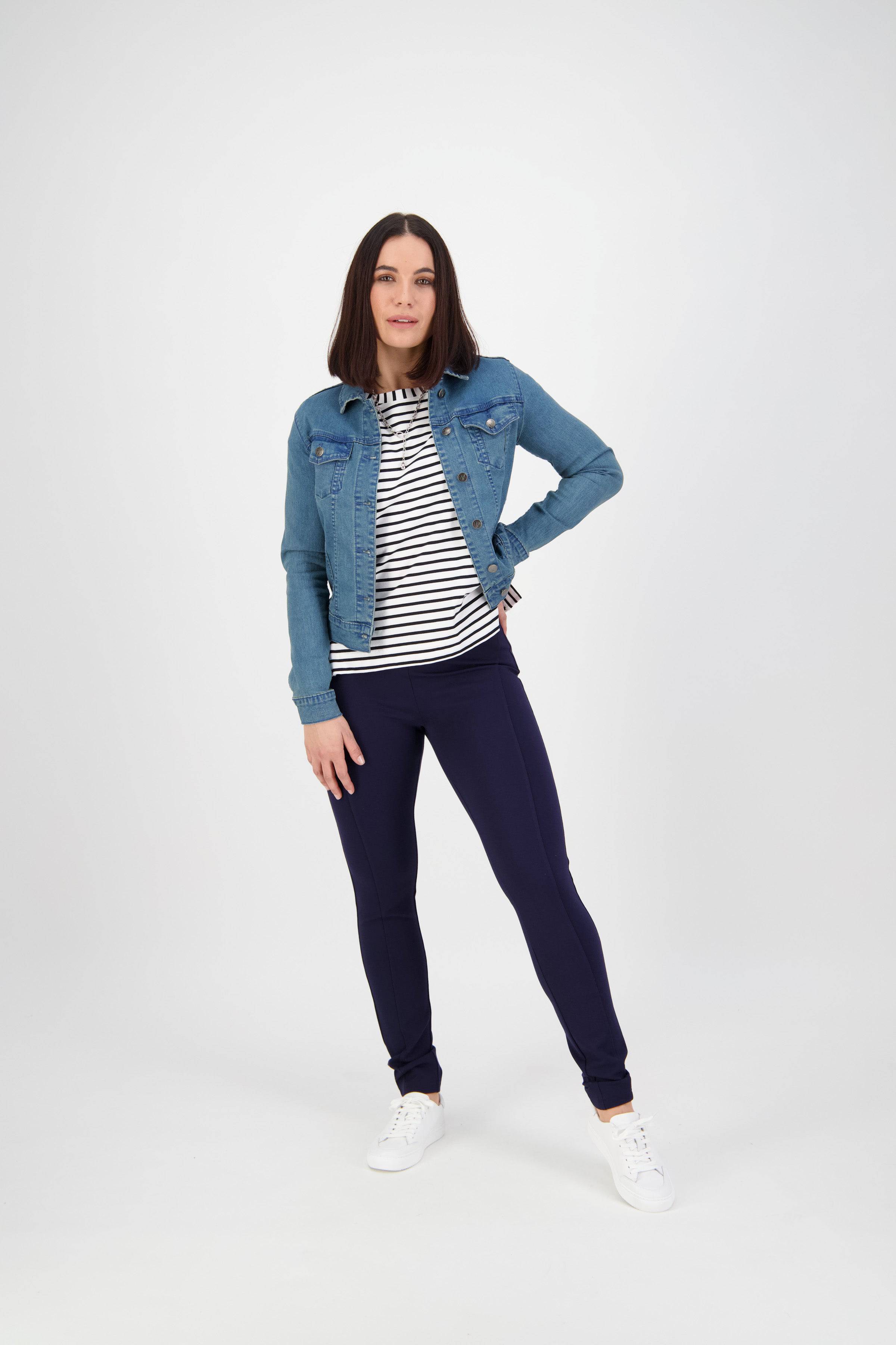 A woman stands confidently against a plain white background, wearing a denim jacket over a black-and-white striped shirt, navy blue pants, and white sneakers.