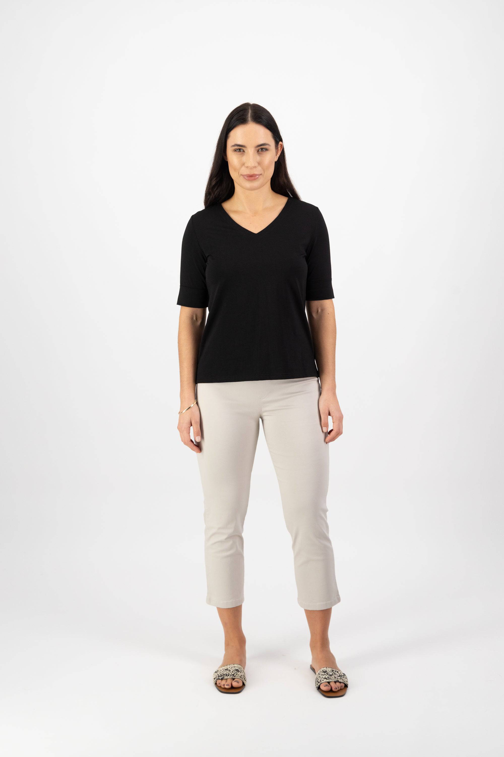 Woman with long dark hair wearing a black V-neck top, light beige cropped pants, and patterned slip-on shoes stands facing forward against a plain white background.