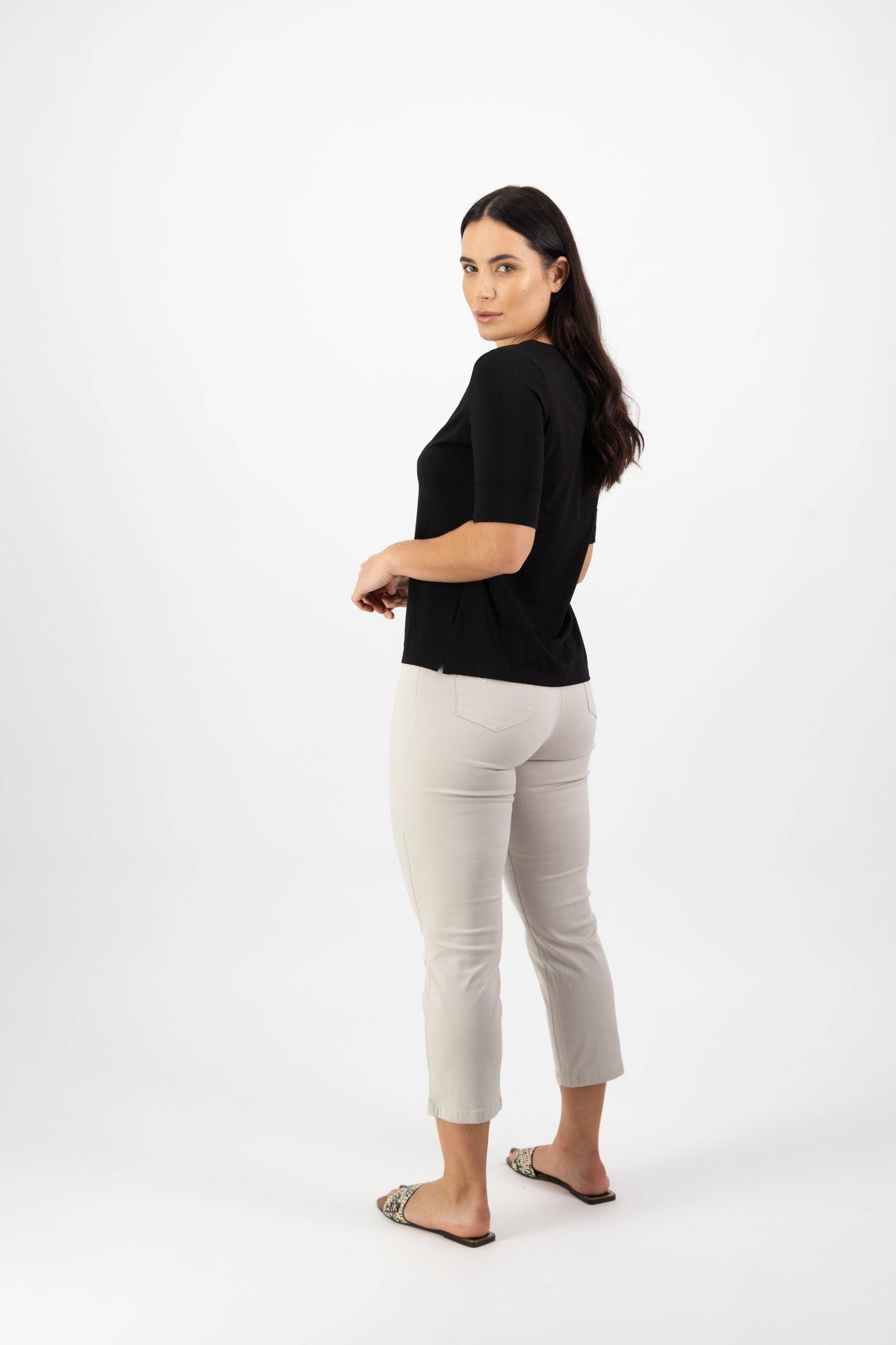 A woman with long dark hair, wearing a black top, light beige pants, and flat shoes, stands on a white background and looks over her shoulder toward the camera.