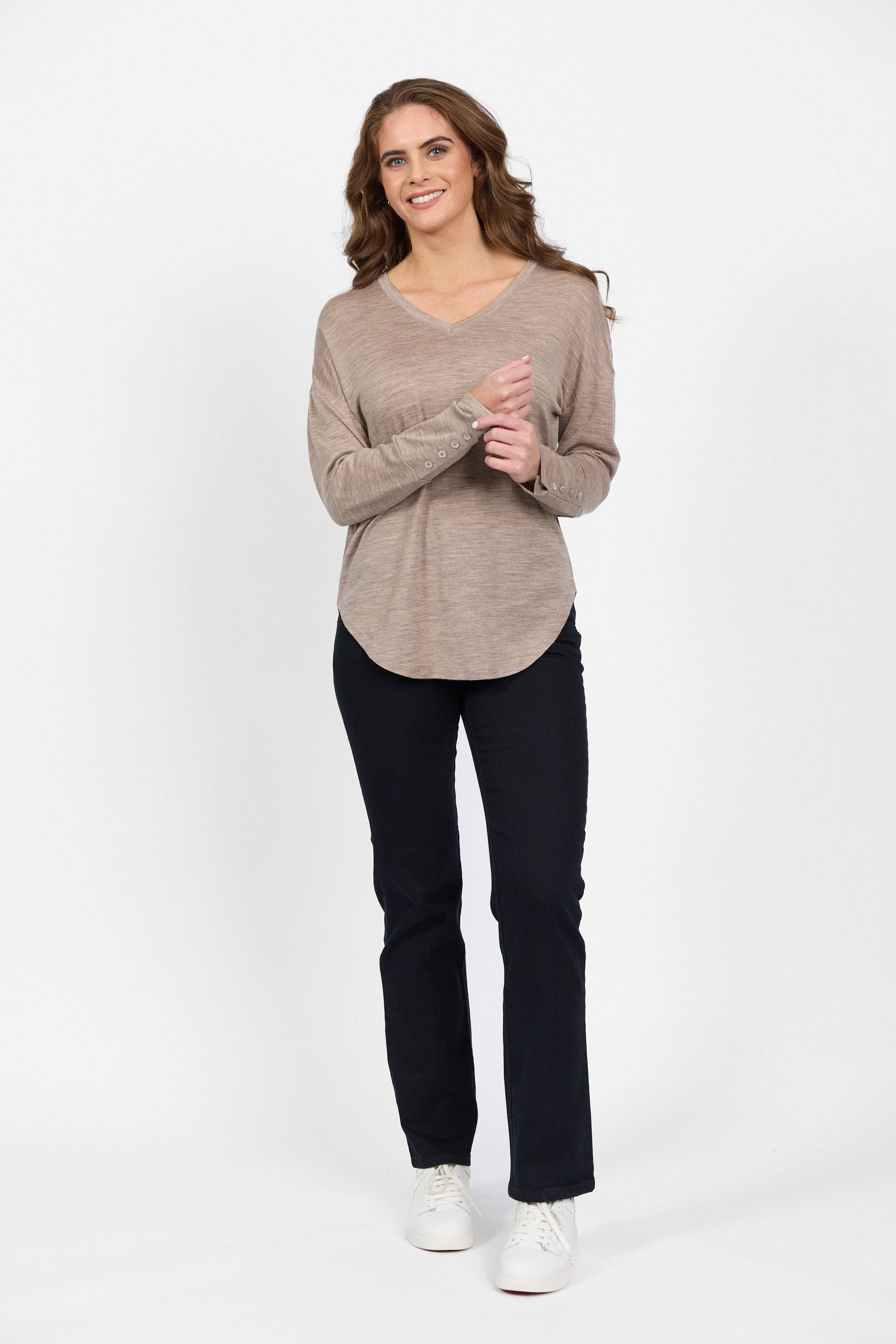 A woman with long brown hair, wearing a beige long-sleeve top, dark pants, and white sneakers, stands smiling against a plain white background.
