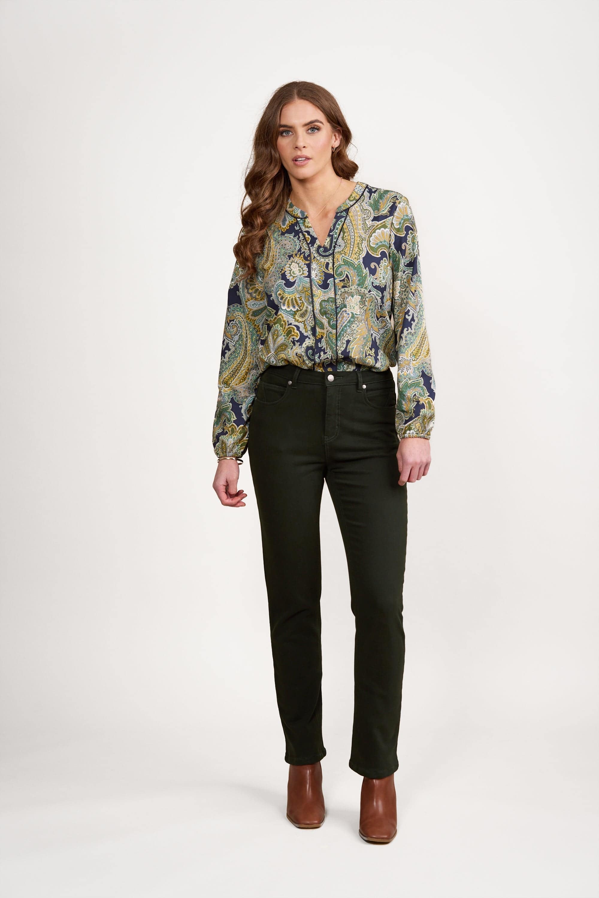 A woman stands against a white background, wearing a patterned blouse with green and blue designs, black pants, and brown ankle boots. She has long wavy hair and a relaxed expression.