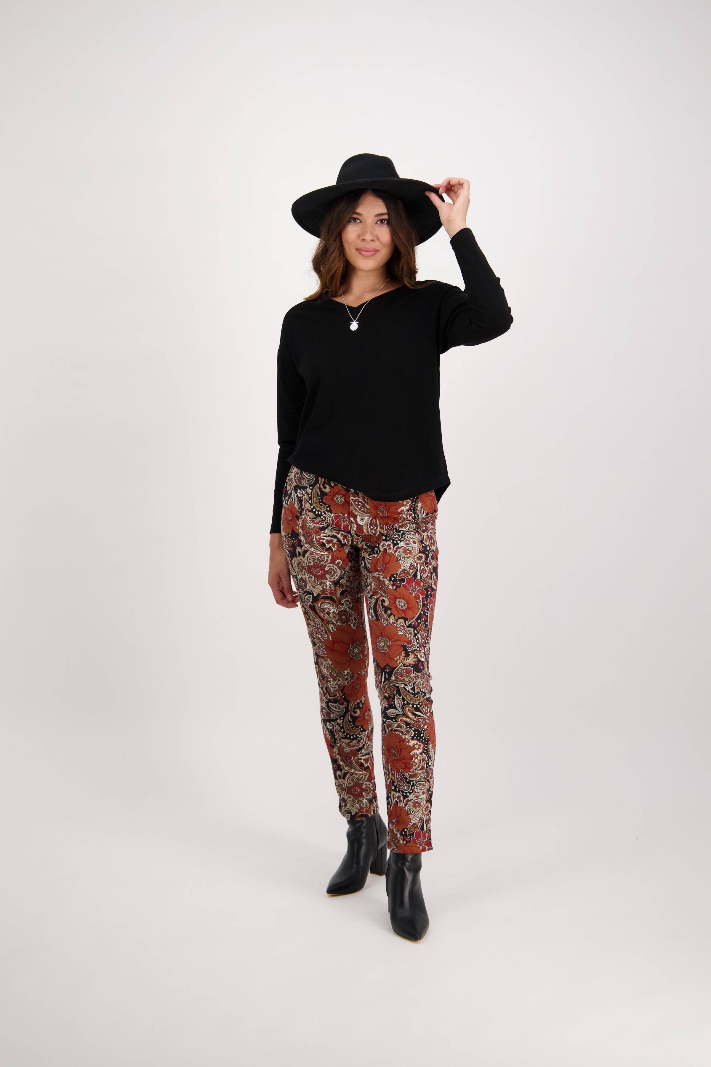Woman standing against a plain background, wearing a black hat, black long-sleeve top, patterned red and brown pants, and black ankle boots, holding her hat with one hand and smiling slightly.