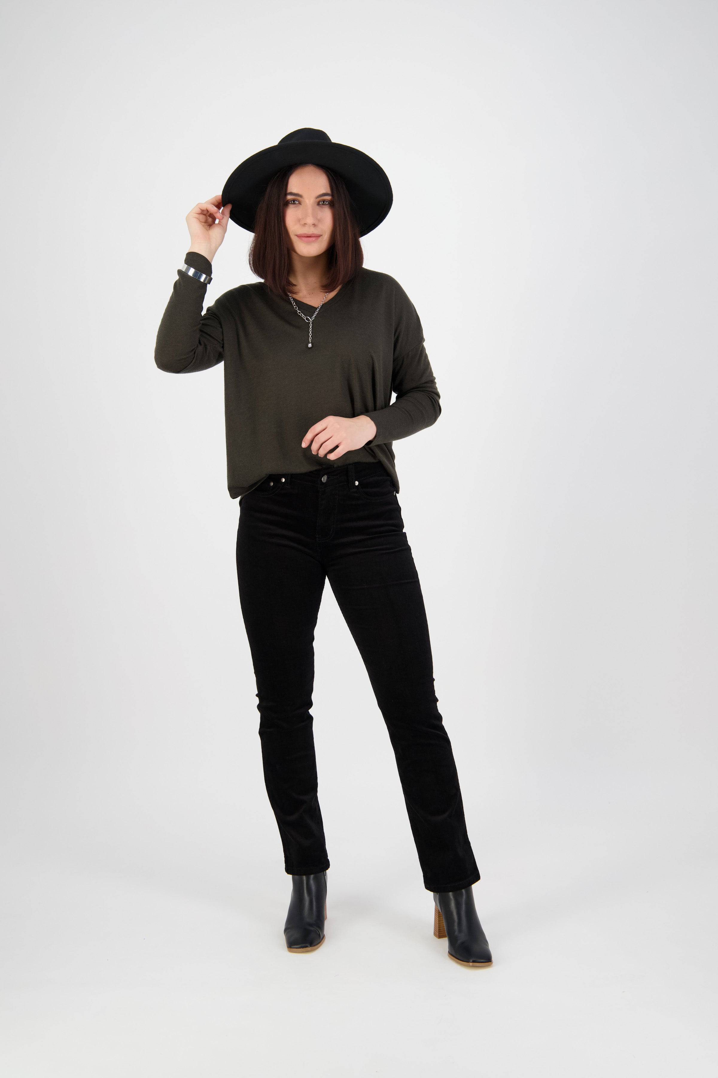 A woman stands against a plain white background, wearing a black wide-brim hat, an olive green long-sleeve top, black pants, and black ankle boots. She holds the brim of her hat with one hand.