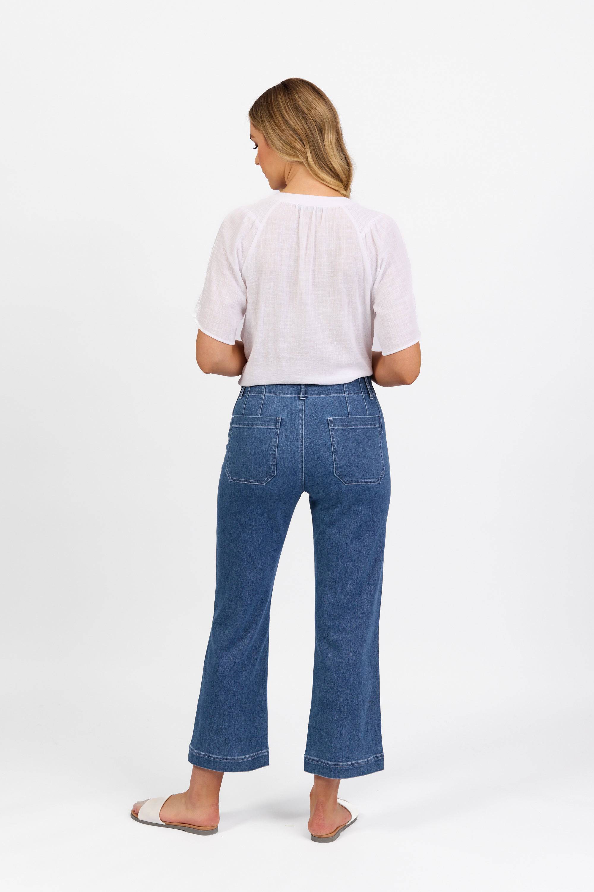 A woman with long blonde hair stands facing away, wearing a white short-sleeve blouse, blue high-waisted jeans with back pockets, and beige slide sandals against a plain white background.