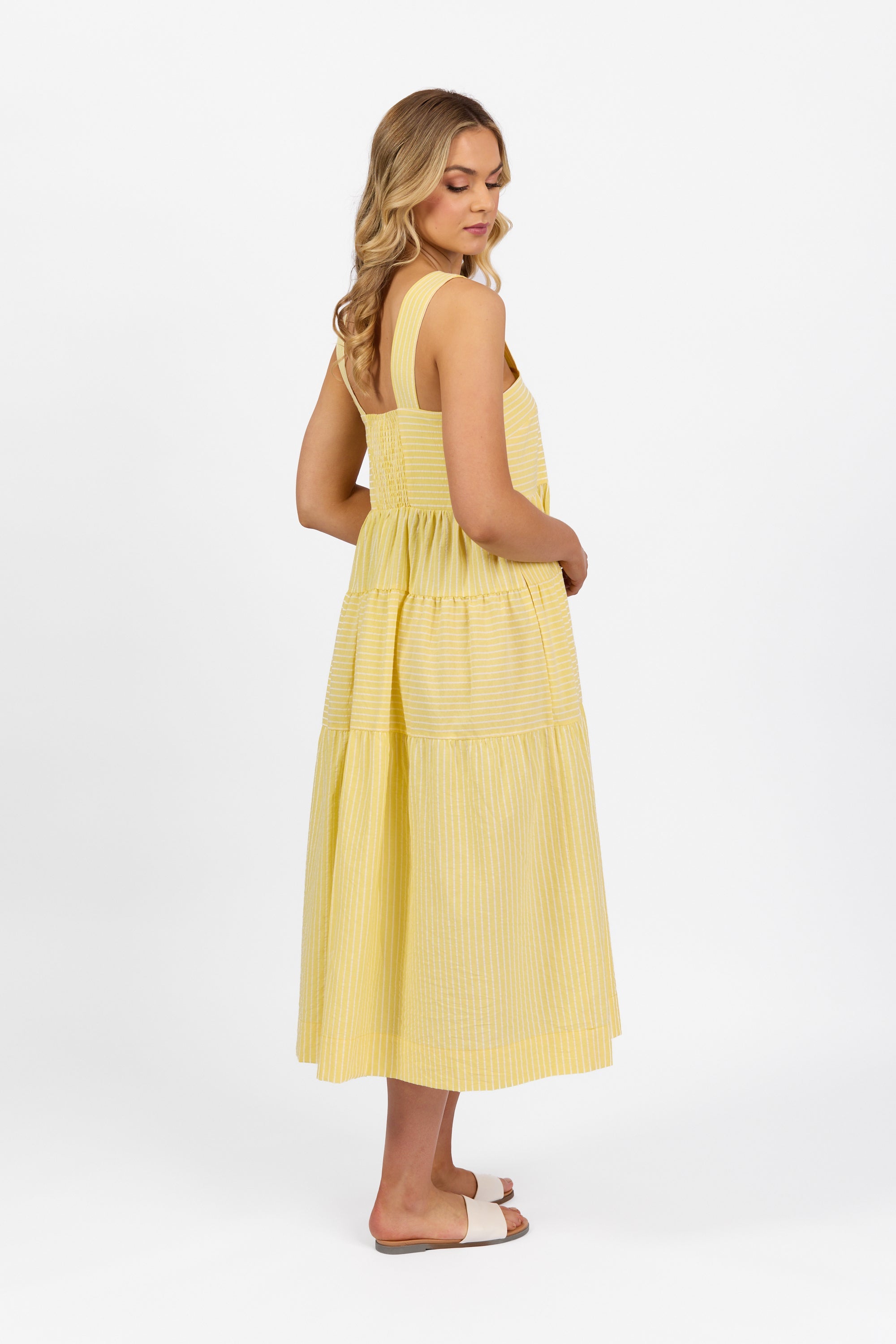 Woman in a yellow, sleeveless, ankle-length dress with wide straps and white stripes stands sideways, looking down, wearing white slide sandals against a plain white background.