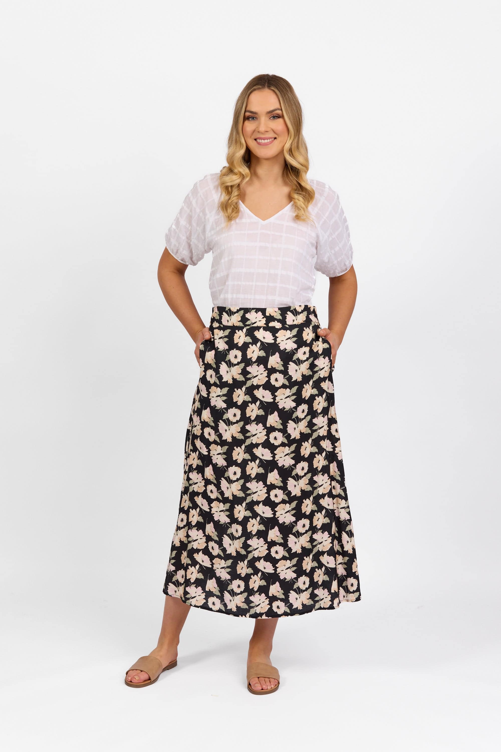 Woman standing and smiling, wearing a white short-sleeve blouse and a black A-line skirt with a white floral pattern. She has long, wavy blonde hair and is wearing tan sandals. Plain white background.