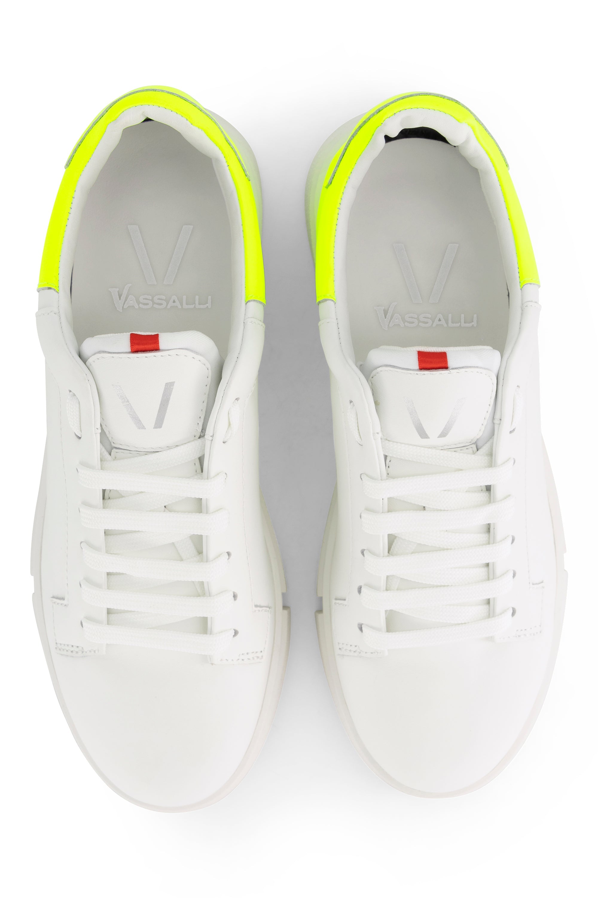 A pair of white Vassalli sneakers with neon yellow accents on the heel and red tabs near the tongue, viewed from above on a white background.