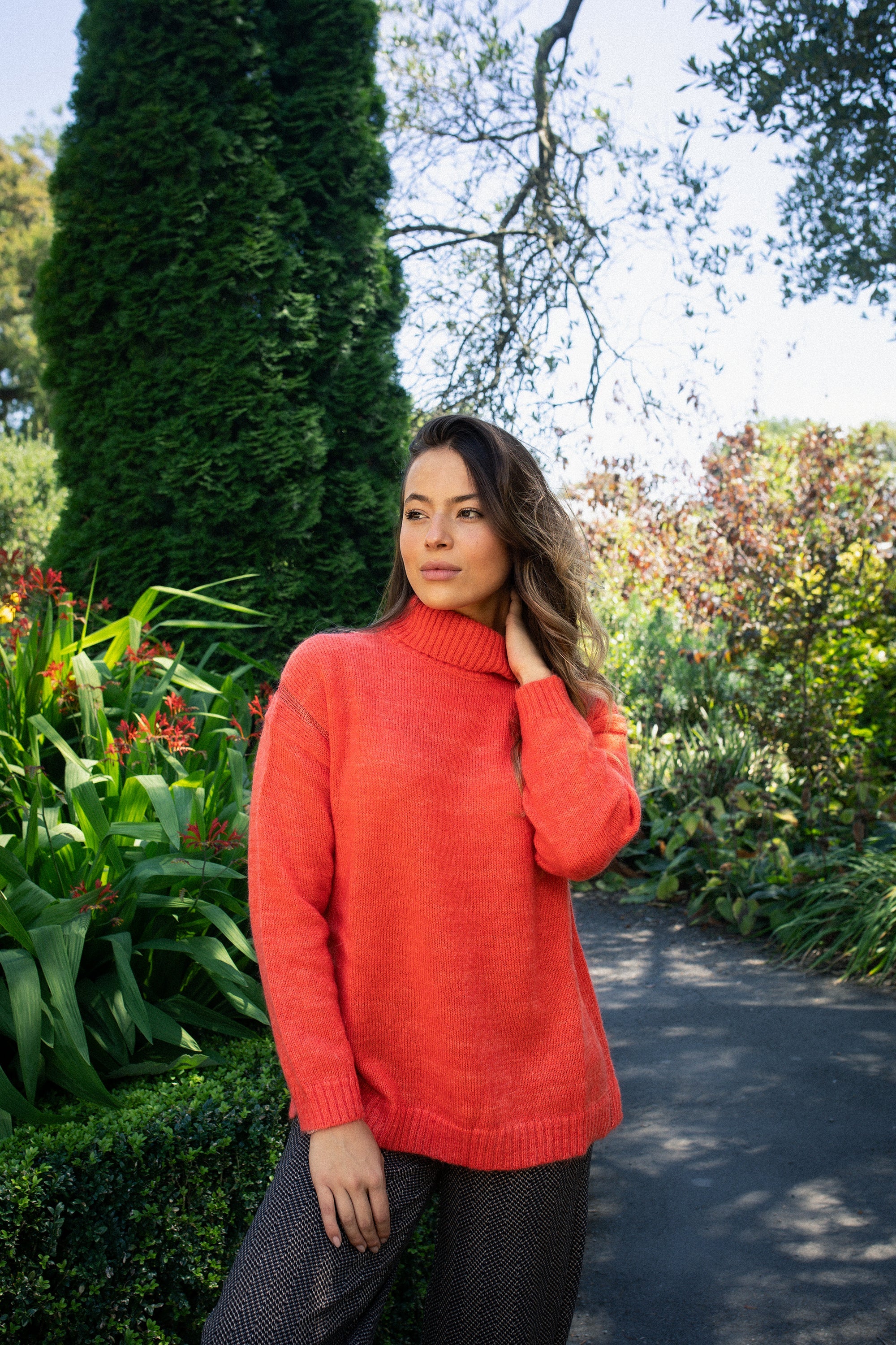 Woman wearing a bright orange sweater and patterned pants stands outdoors in a lush, green garden, touching her neck and looking thoughtfully into the distance.