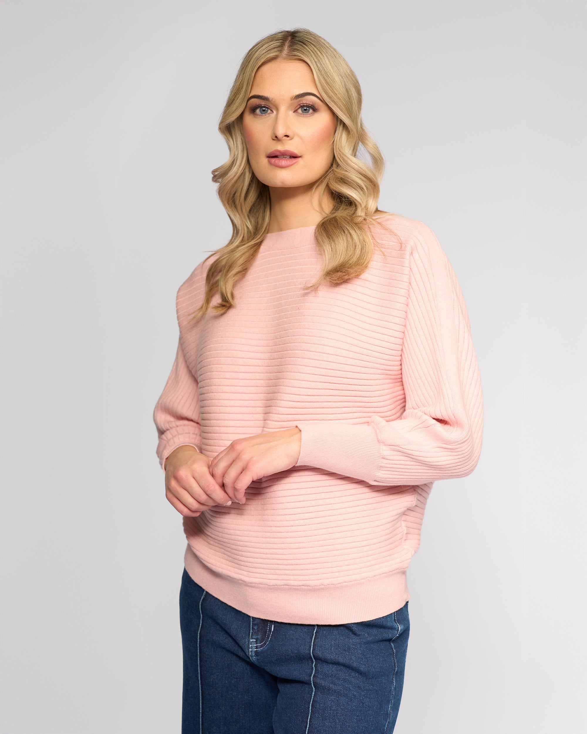 A woman with long blonde hair wearing a light pink ribbed sweater and blue jeans stands against a plain light gray background, looking at the camera with a neutral expression.