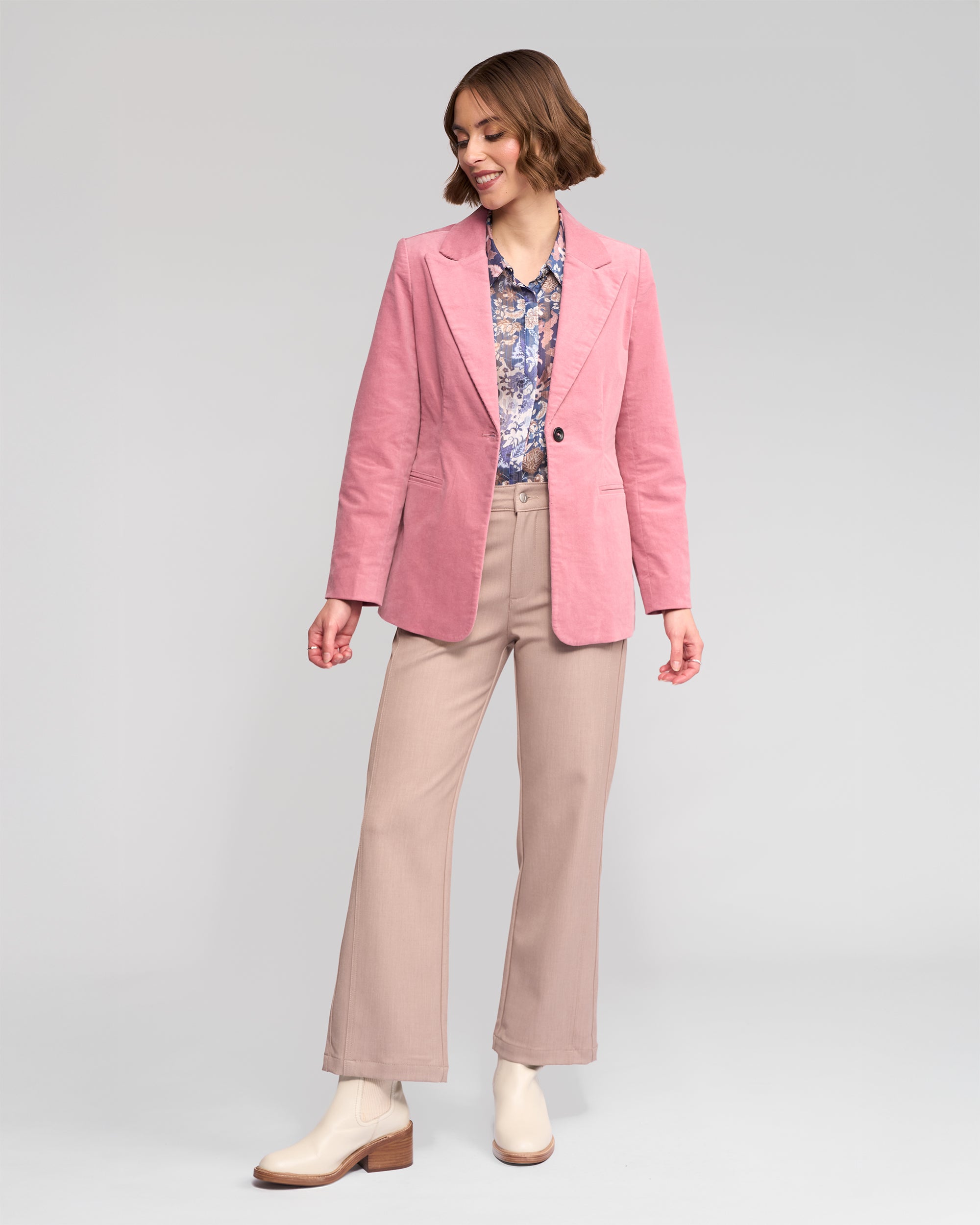 A woman is standing and smiling, wearing a pink blazer, floral shirt, beige pants, and white ankle boots, on a plain light gray background.