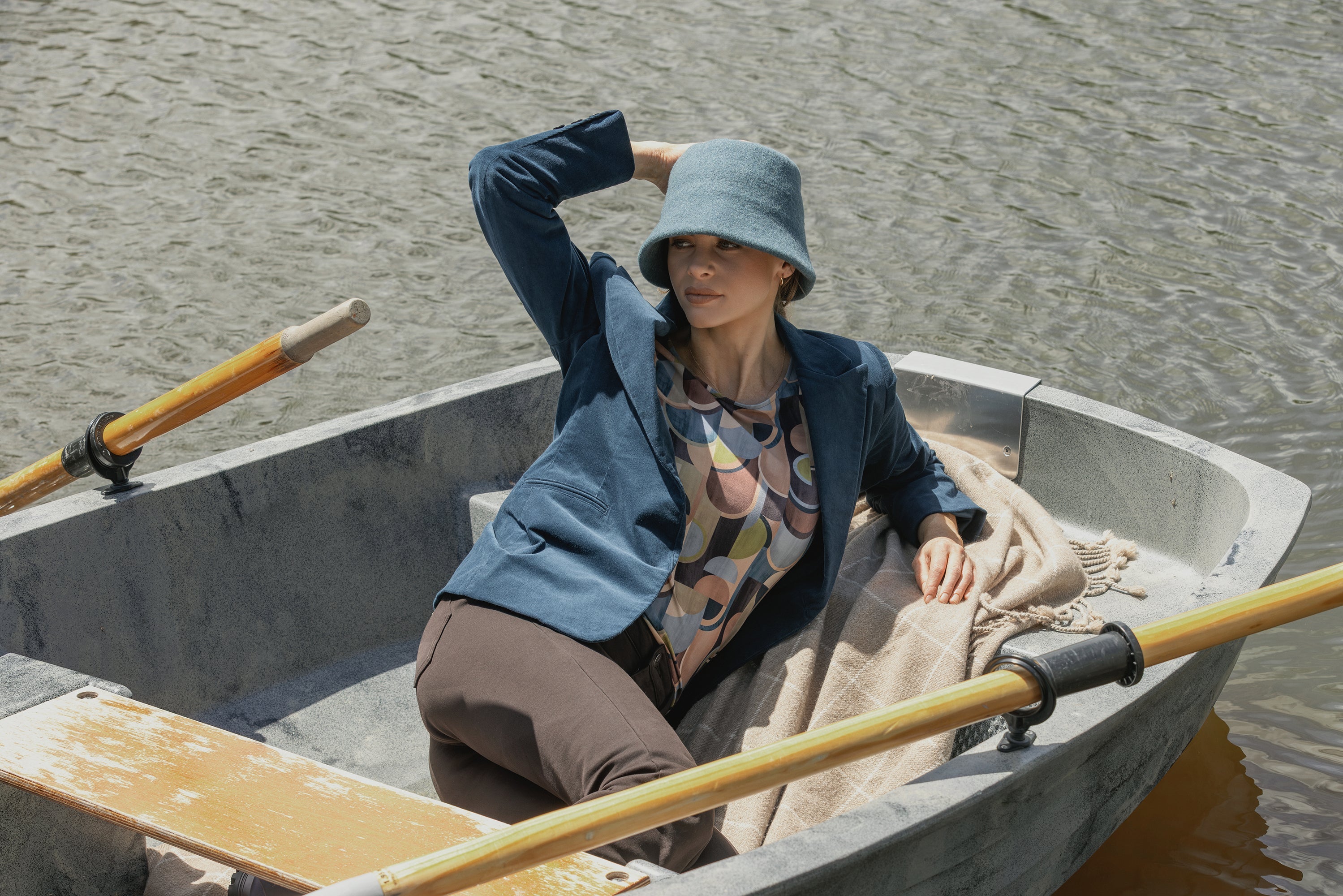 A woman in a blue hat and blazer sits relaxed in a rowboat on the water, with one hand holding her hat and a blanket beside her. Oars rest on either side of the boat.