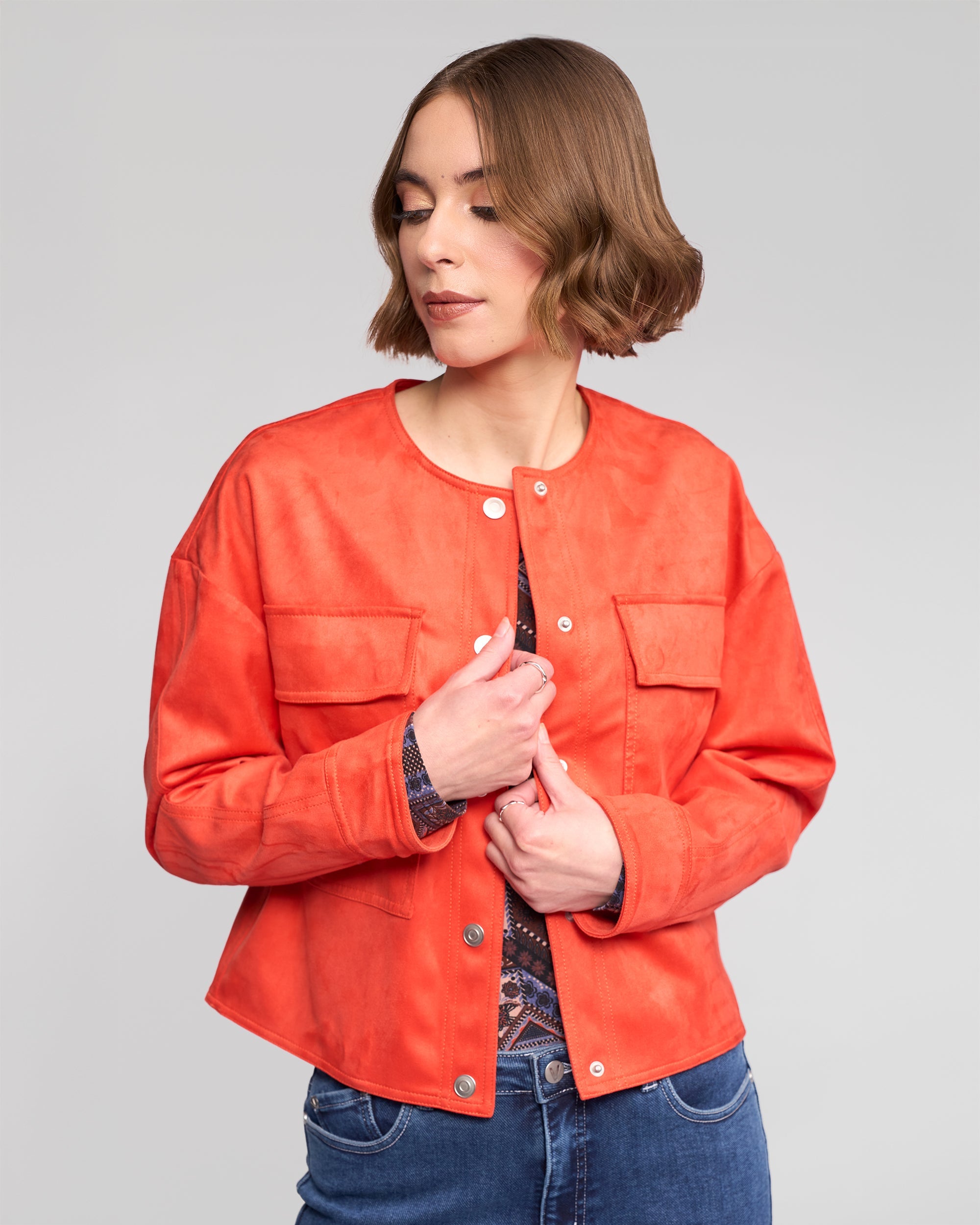 A woman with short brown hair wears an orange button-up jacket over a patterned top and blue jeans, looking down with her hands adjusting the jacket. The background is plain light gray.