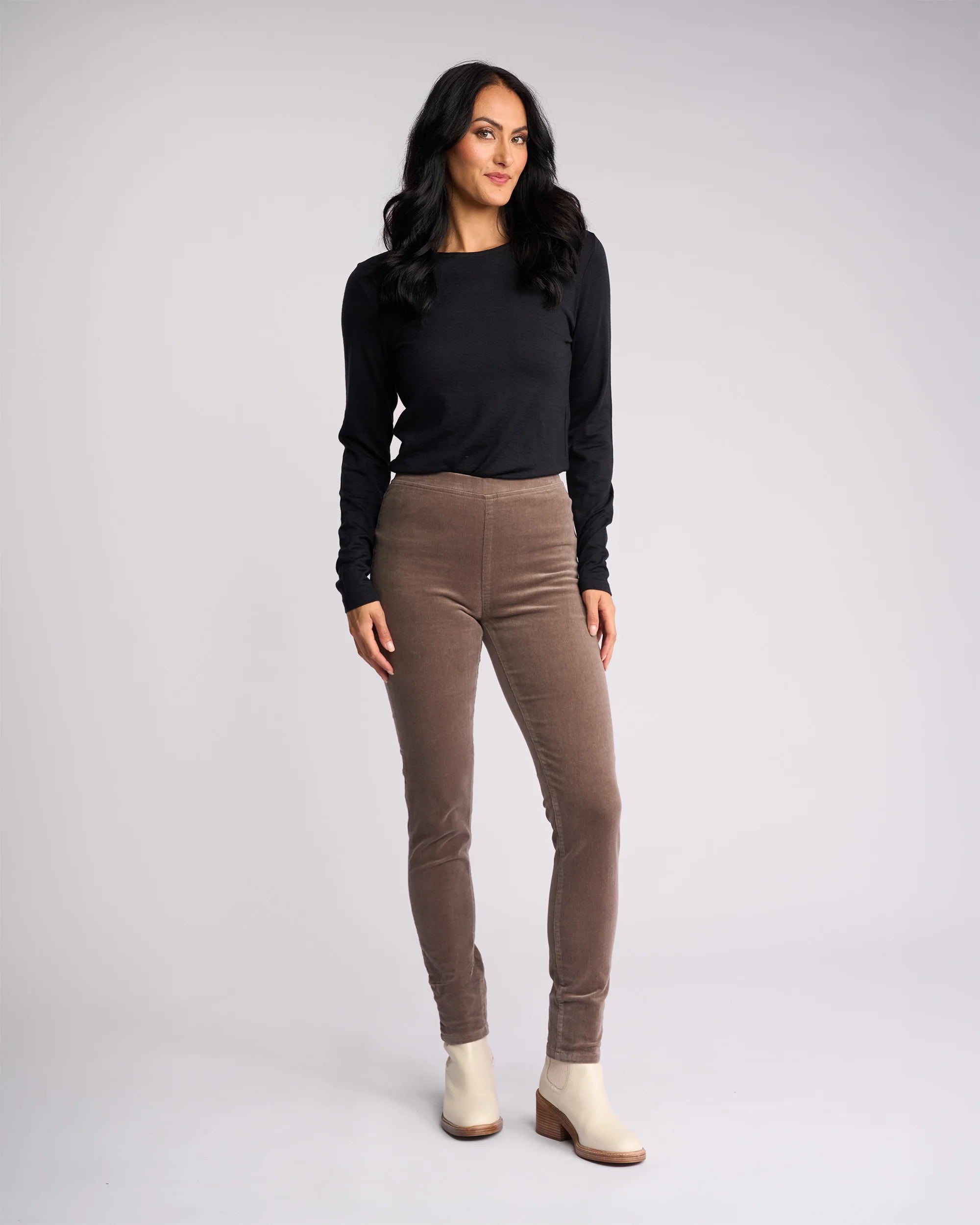 A woman with long dark hair wears the Vassalli Origin Cord Pull On in taupe, paired with a black long-sleeve top and cream ankle boots. She stands against a light gray background, smiling slightly with one hand by her side and the other bent.
