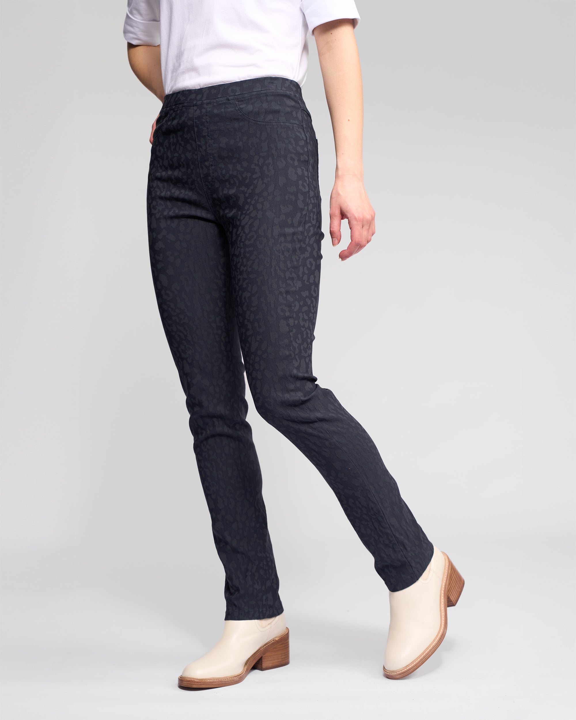 A person wearing black patterned slim-fit pants, a white top with rolled sleeves, and light-colored ankle boots stands against a plain light gray background.