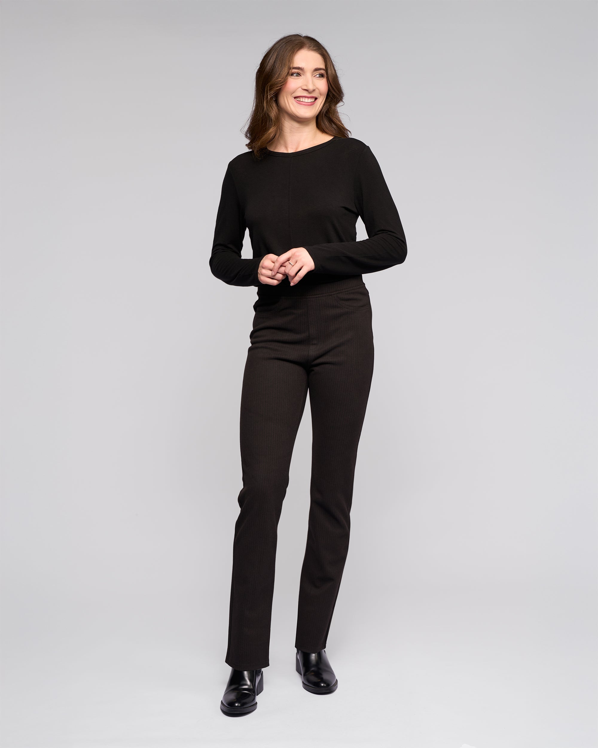 Woman with wavy brown hair stands smiling against a plain light gray background, wearing a long-sleeved black top, black pants, and black ankle boots.