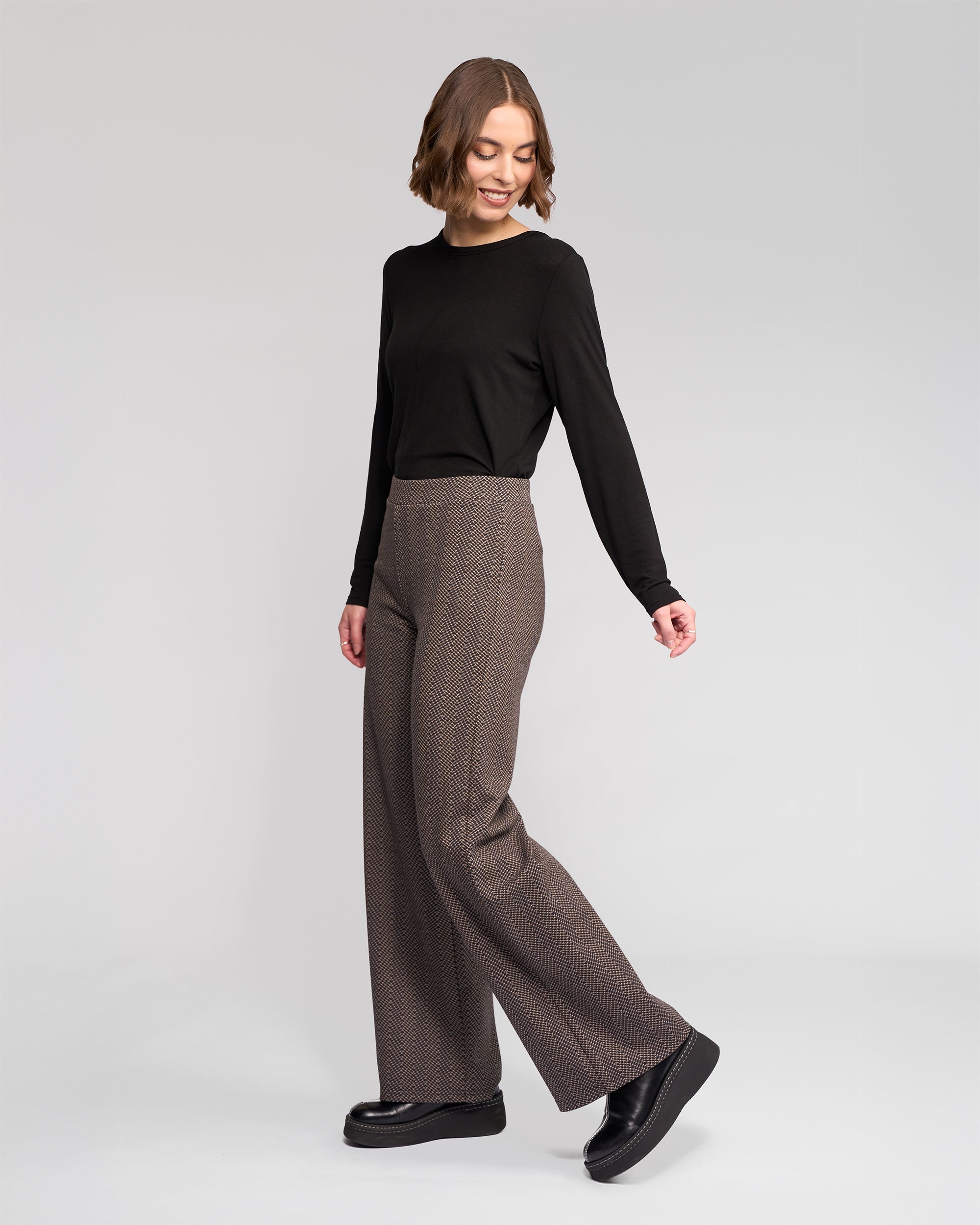 A woman with short brown hair smiles and looks over her shoulder, wearing a black long-sleeve top, brown wide-leg trousers, and black platform shoes, standing against a plain light background.