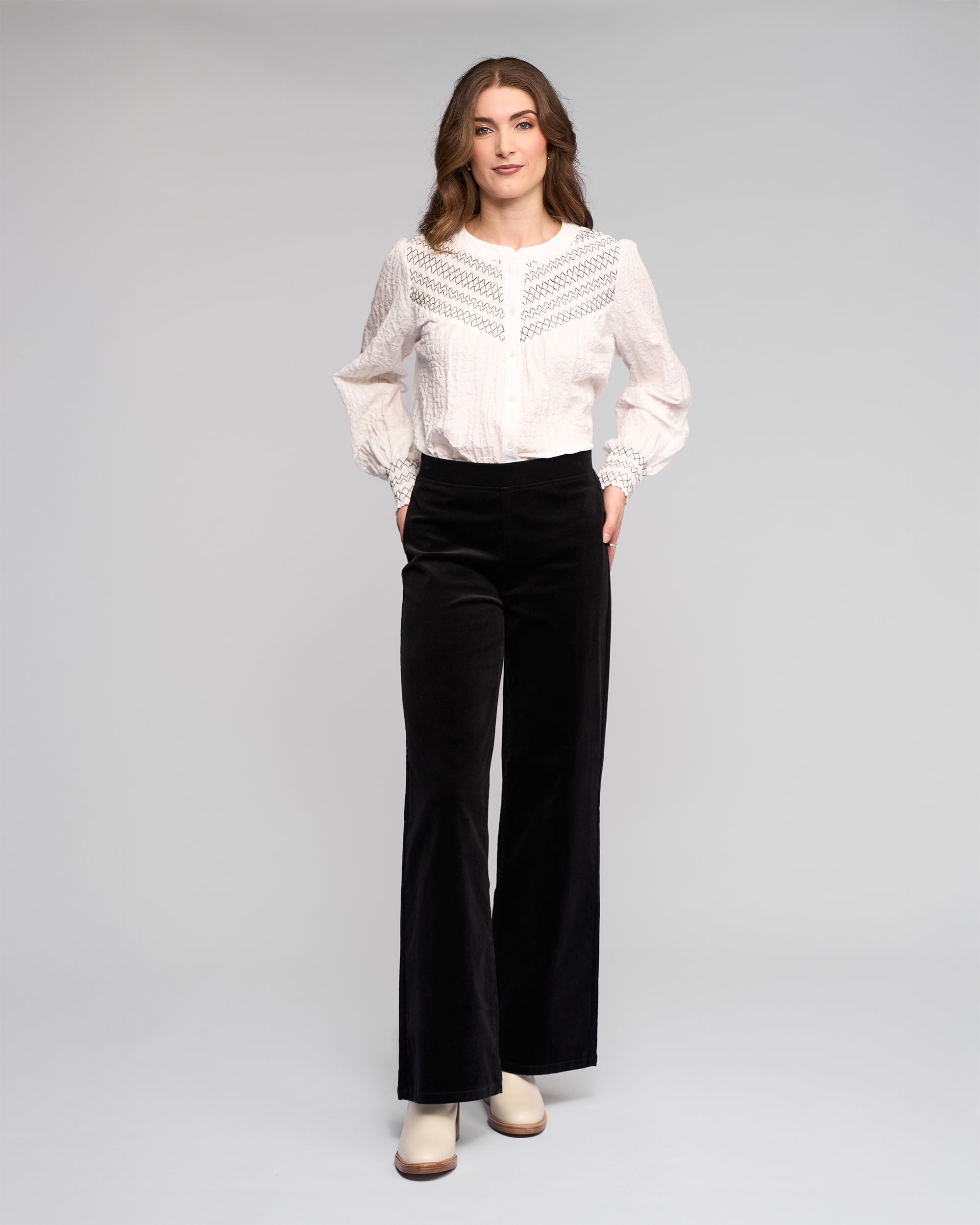 A woman stands against a plain background wearing a white long-sleeve blouse with lace details, black wide-leg pants, and white shoes.