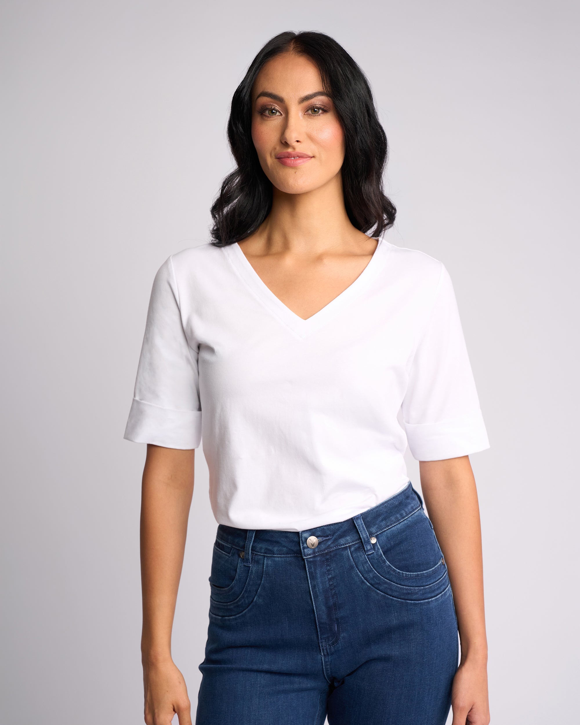 A woman with long dark hair wears the Vassalli Reece Top in white—a V-neck stretch T-shirt—paired with high-waisted blue jeans, standing against a light gray background and looking forward with a slight smile.
