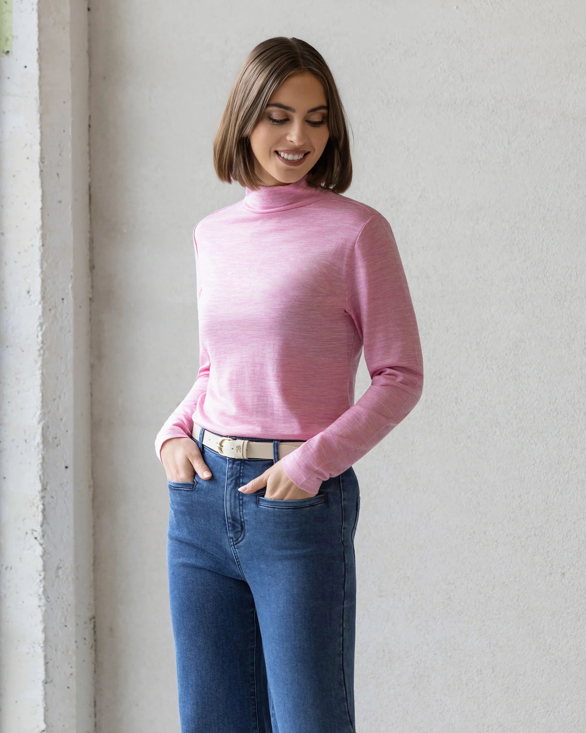 A woman with a bob haircut wears a light pink long-sleeve mock-neck top and blue jeans, standing against a neutral wall with her hands in her pockets, smiling and looking down.