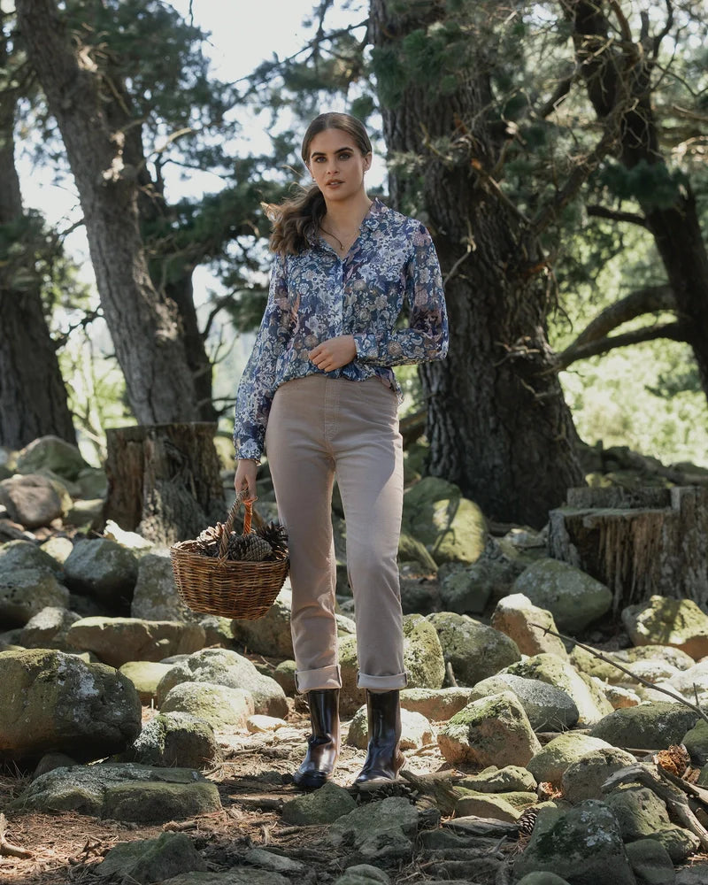 Young woman with short brown hair stands against a plain background, wearing a long-sleeve patterned top, olive green wide-leg pants, and black shoes, with arms folded and head slightly tilted.
