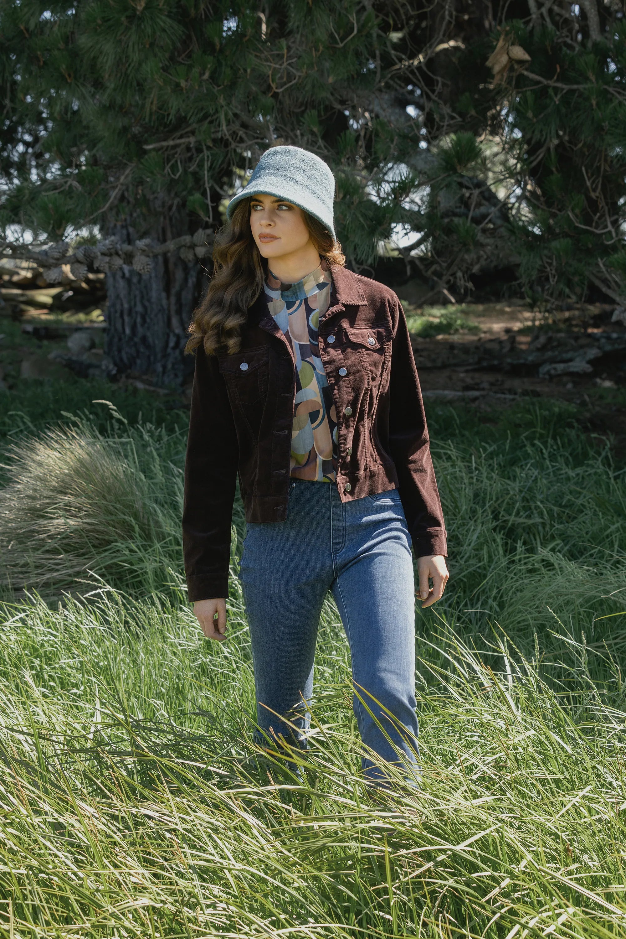 A woman wearing a light gray bucket hat, dark brown jacket, patterned blouse, and blue jeans stands in tall green grass with trees in the background.