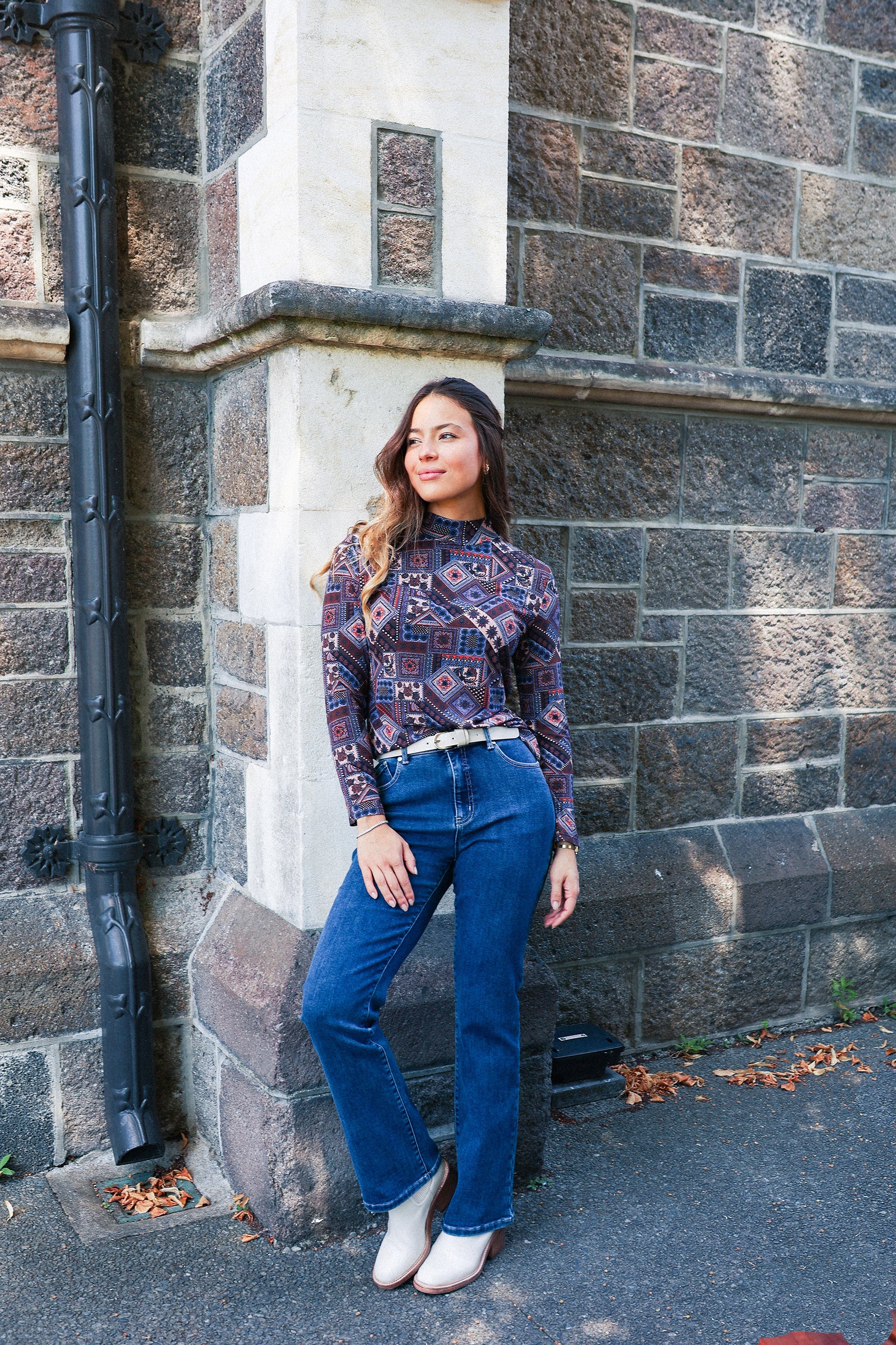 A woman in a patterned long-sleeve top, blue jeans, and white shoes stands against a stone wall, smiling with one hand resting on the wall and the other by her side. Dry leaves are scattered on the ground.