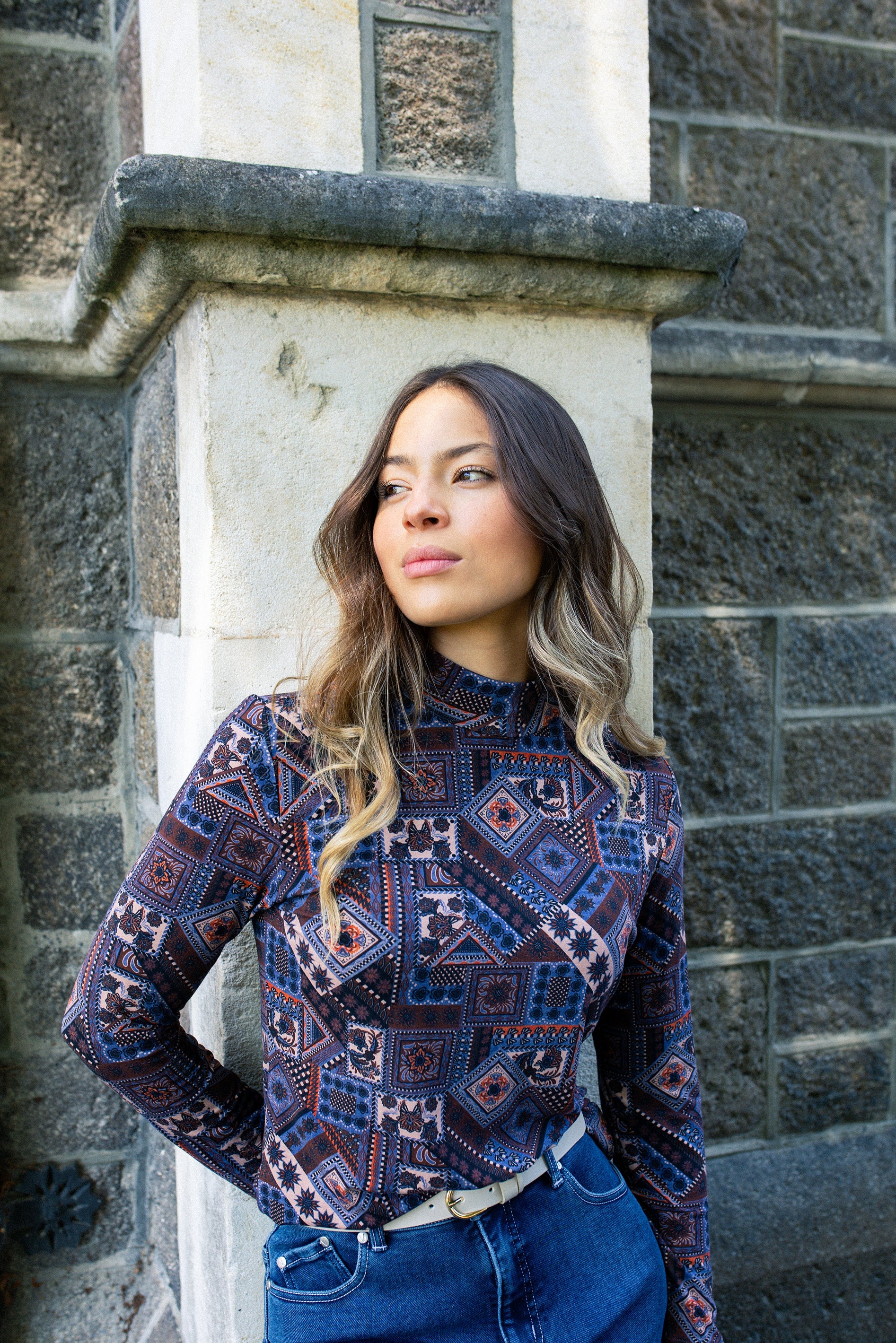 A woman with wavy hair wearing a patterned turtleneck and blue jeans stands against a stone wall, looking to the side with a thoughtful expression.