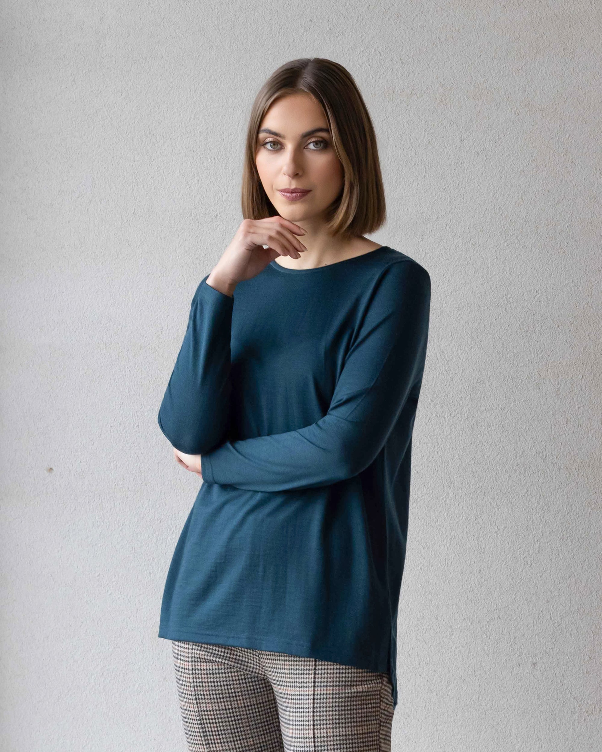A woman with straight brown hair in a bob, wearing a long-sleeved teal top and plaid pants, stands against a light-colored wall, resting her chin on her hand and looking at the camera.