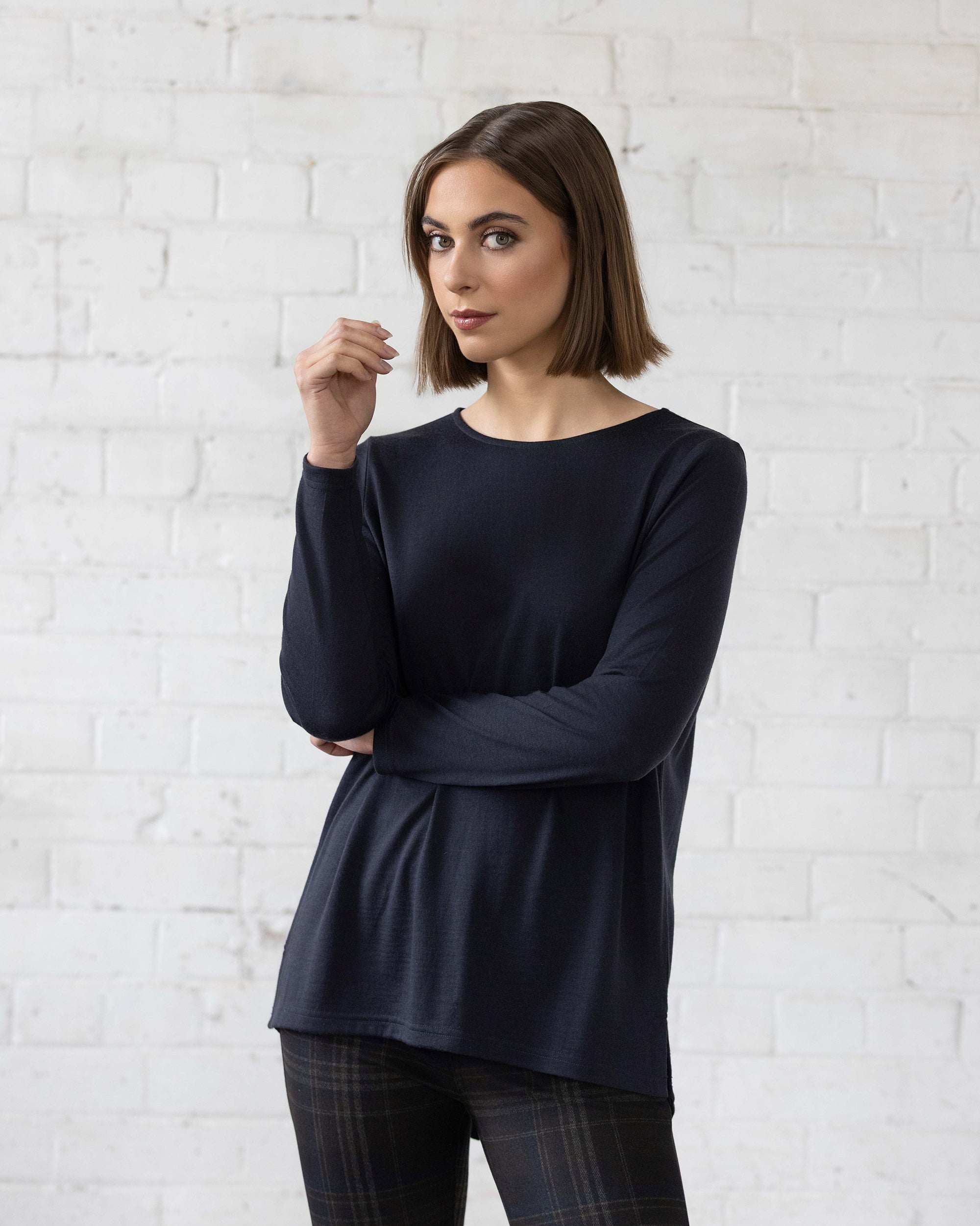 A woman with straight brown hair wearing a long-sleeved navy top and plaid pants stands in front of a white brick wall, looking at the camera with her arms crossed.