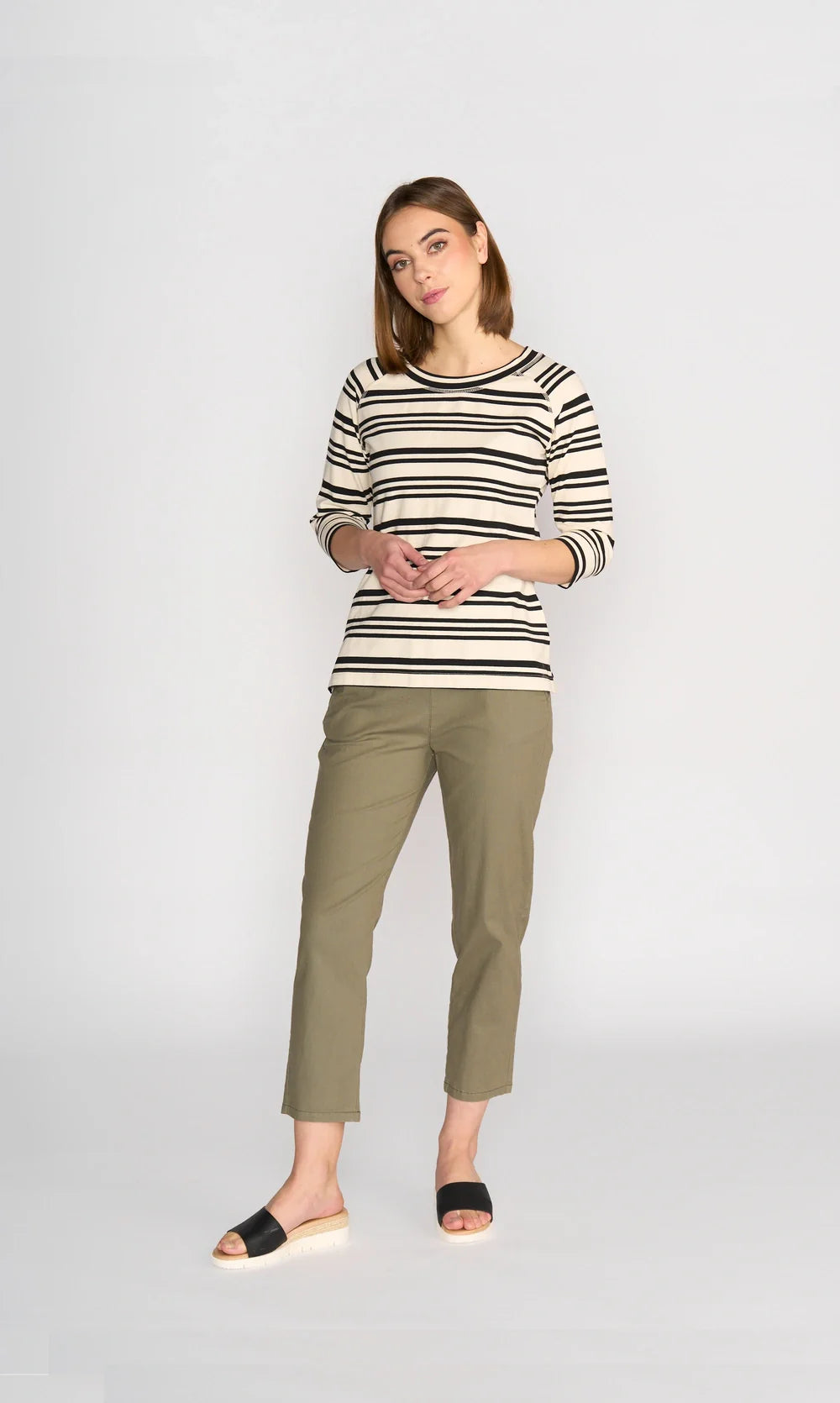 A woman with wavy blonde hair wears a light blue long-sleeve top, beige wide-leg pants, and off-white ankle boots, standing against a plain light gray background.