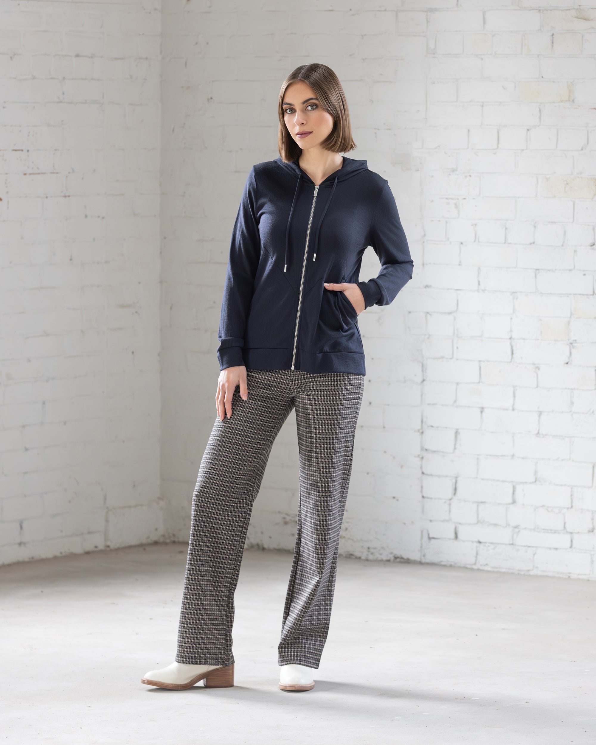A woman stands in a bright, minimalist room wearing a navy zip-up hoodie, gray plaid pants, and white shoes. She faces forward with one hand in her pocket and a relaxed expression.