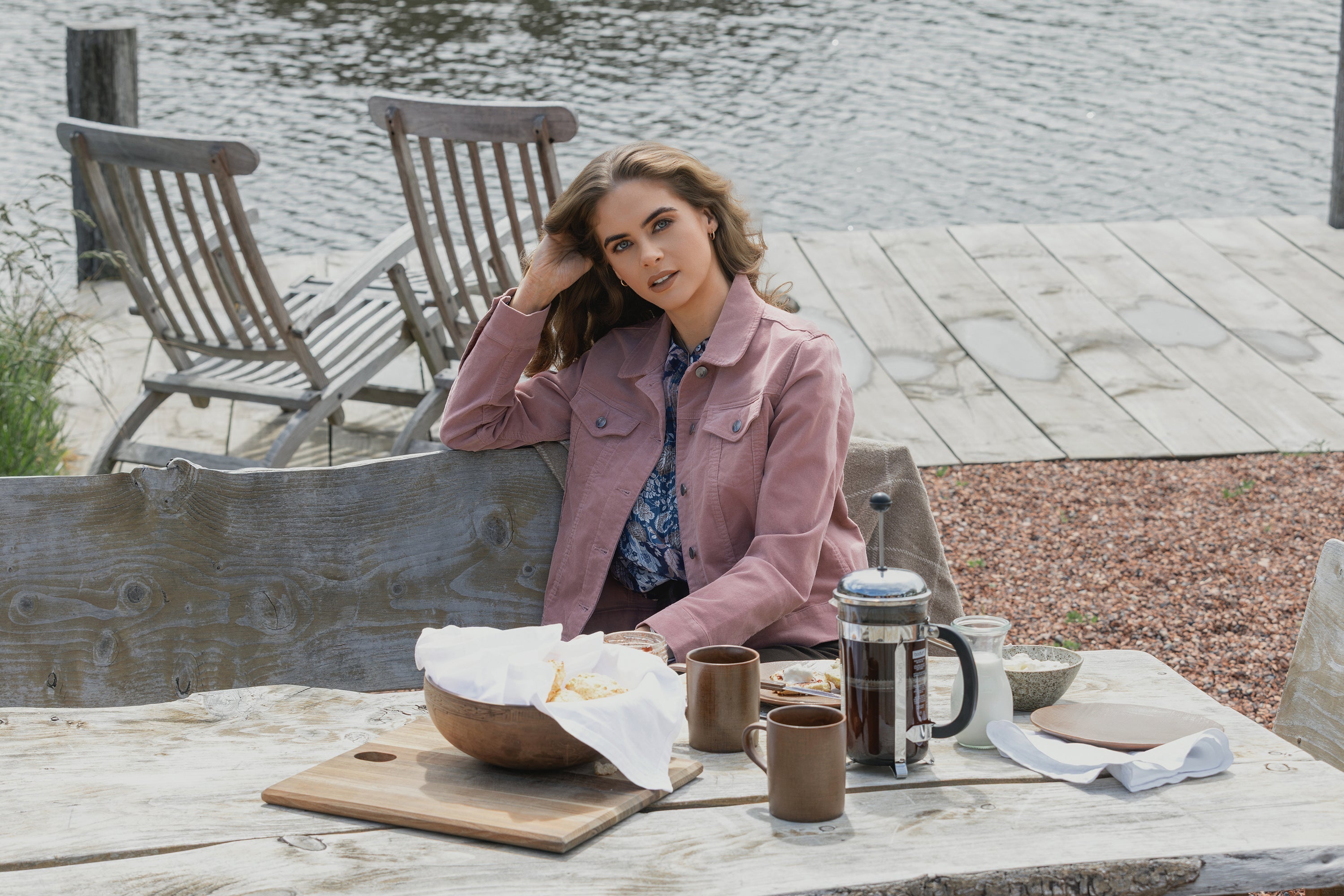 A woman in a pink jacket sits at a rustic outdoor table by a lake, with coffee, cups, and a bowl of bread in front of her. Wooden chairs and a dock are in the background.
