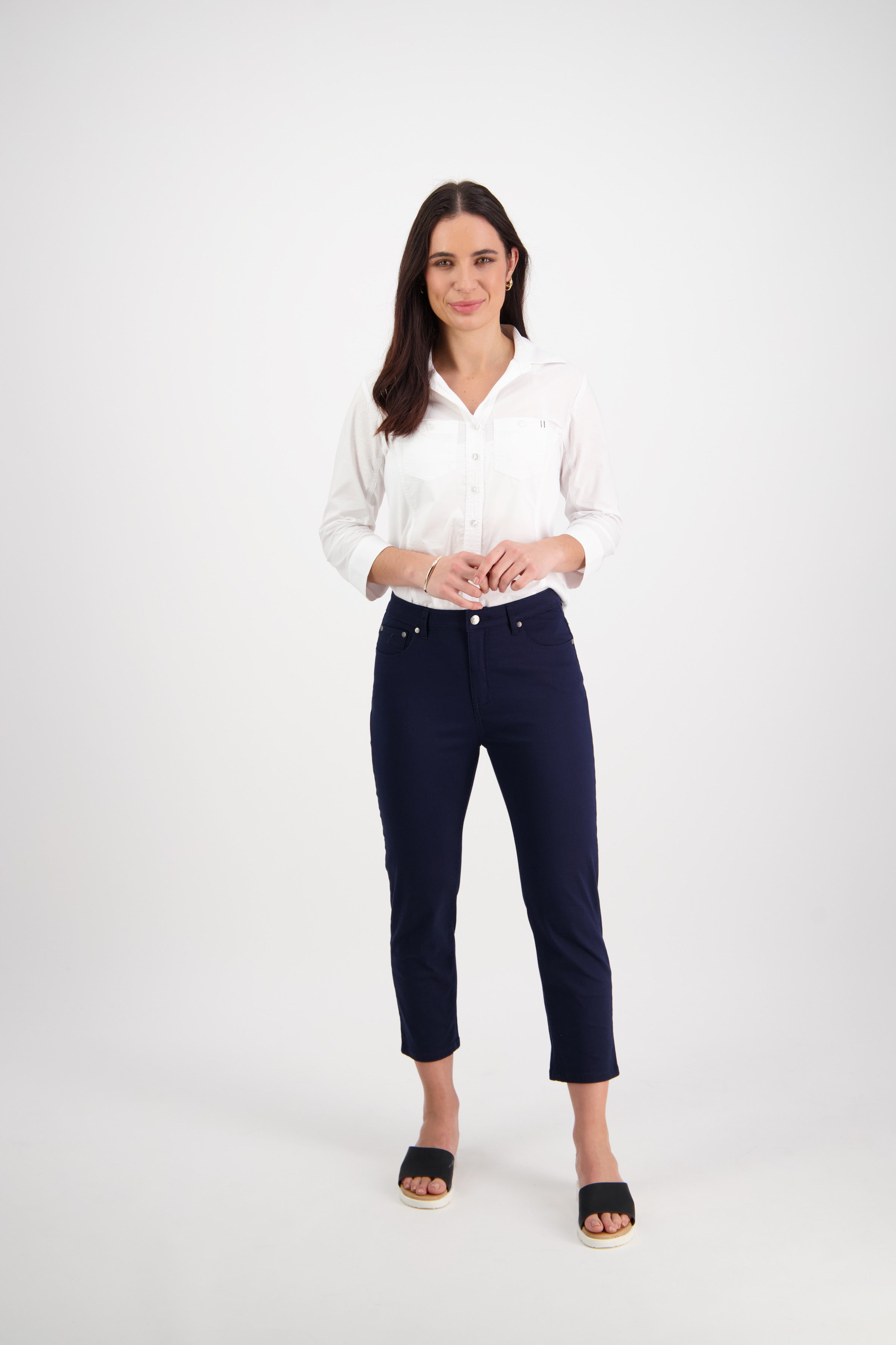 A woman stands against a plain white background, wearing a white button-up shirt, dark navy ankle-length pants, and black slide sandals. She has long dark hair and is smiling slightly.