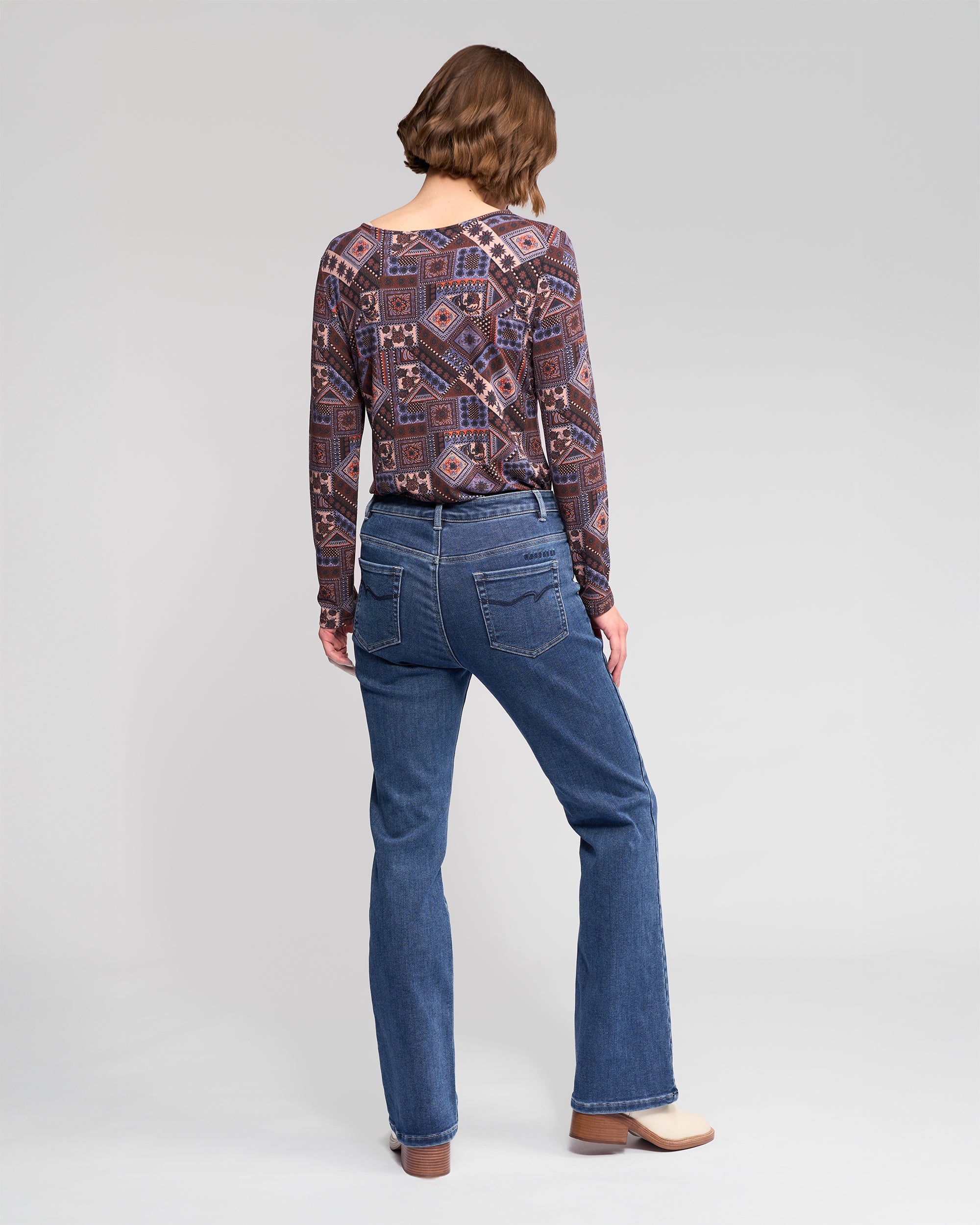 A person with short, wavy brown hair is standing facing away, wearing a long-sleeve patterned shirt, blue jeans, and beige shoes against a plain light gray background.