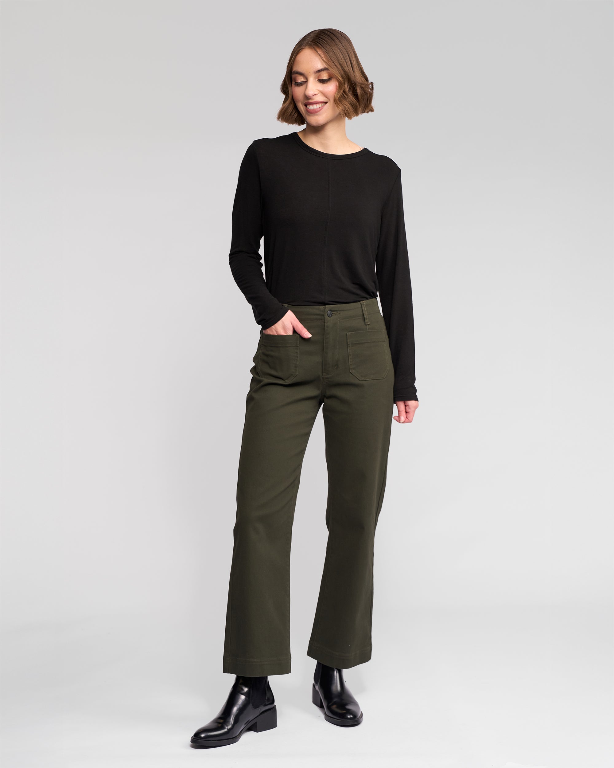 A person stands against a plain light gray background, wearing a long-sleeve black top, olive green high-waisted pants, and black ankle boots, with one hand in their pocket and smiling.