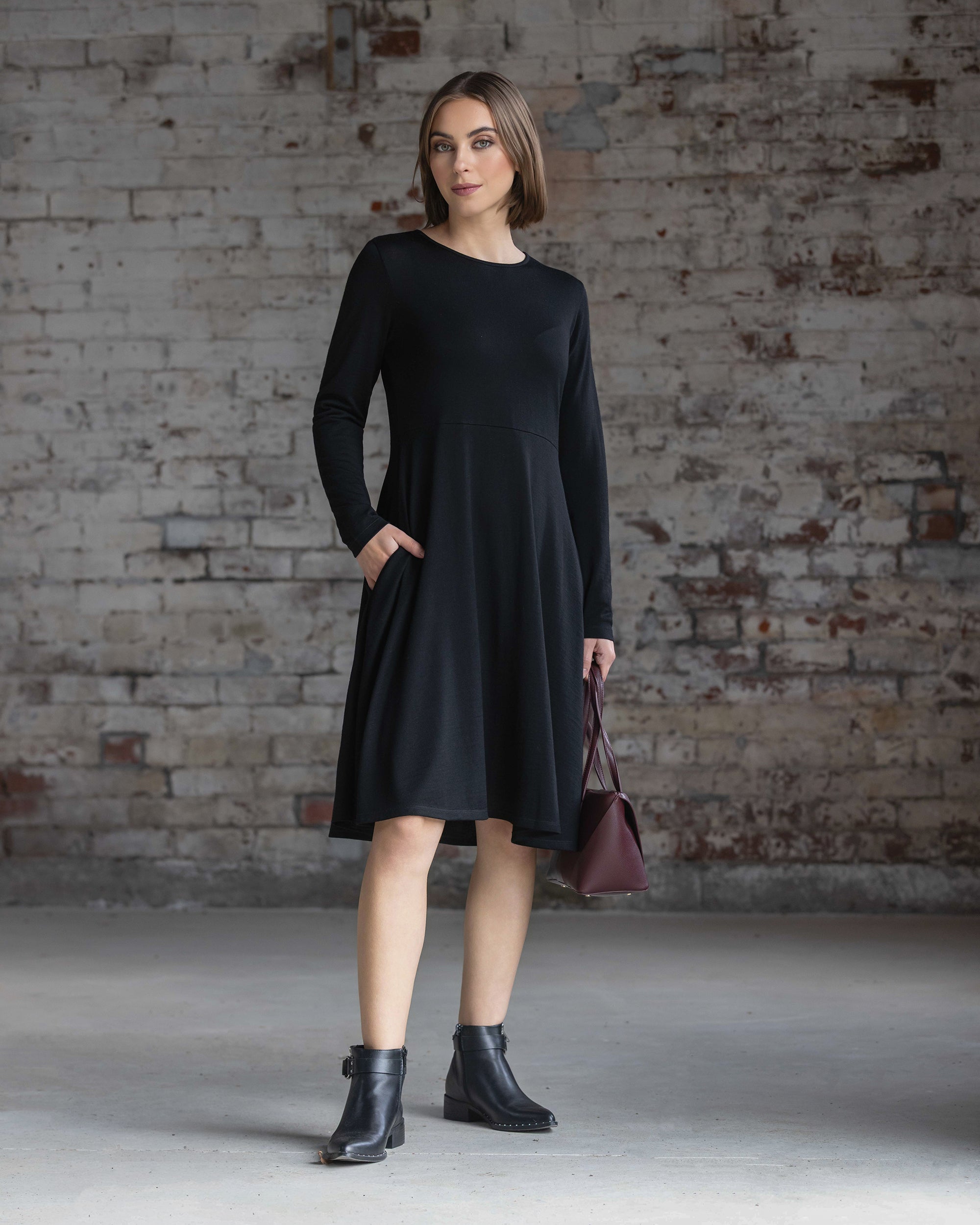 A woman in a long-sleeved black dress and black ankle boots stands in front of a worn brick wall, holding a dark red handbag in one hand and looking slightly to the side.