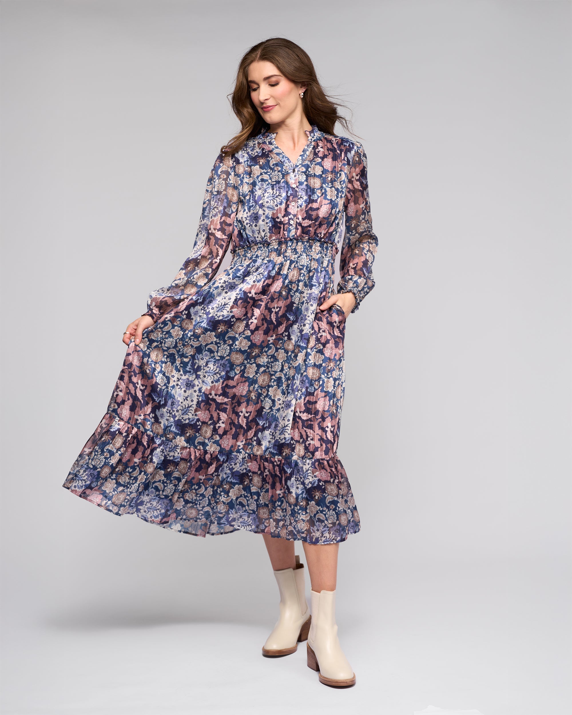 Woman wearing a long-sleeve, floral-patterned midi dress with a ruffled hem, posing against a plain light background, and pairing the dress with cream ankle boots.
