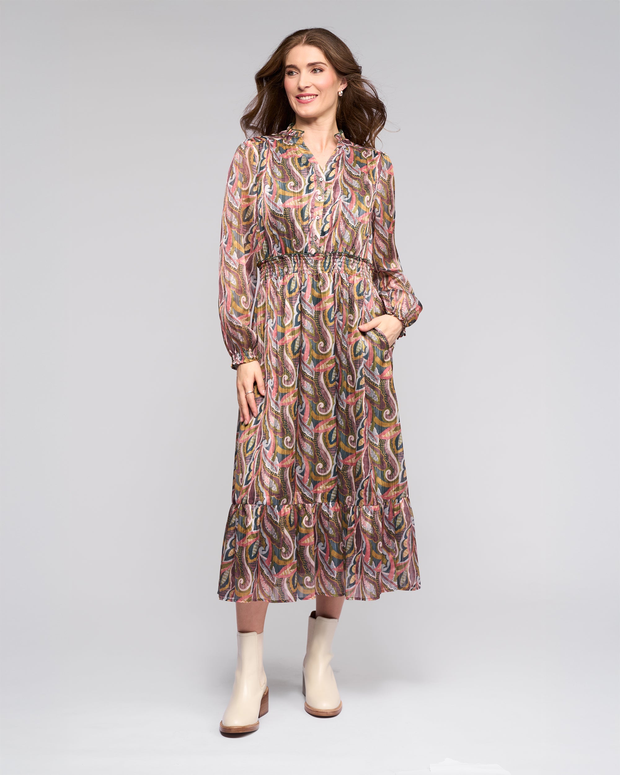 A woman stands against a plain background wearing a long-sleeved, patterned midi dress with a ruffled hem, and light ankle boots. She has wavy brown hair and is smiling with her hand in a pocket.