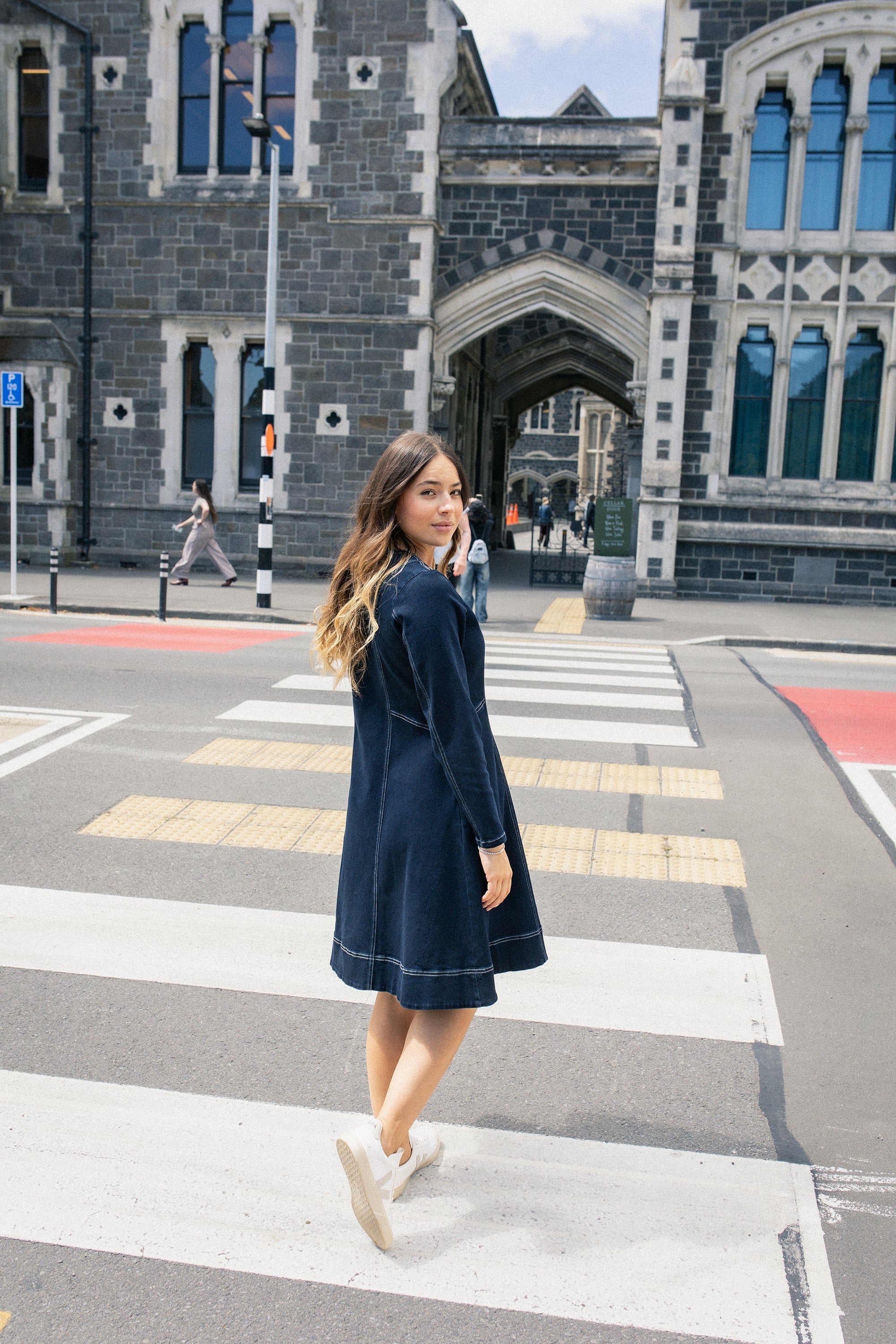 A woman in a navy dress and white sneakers stands on a crosswalk, turning to smile at the camera. Behind her is a historic stone building with arched windows and a cloudy sky.