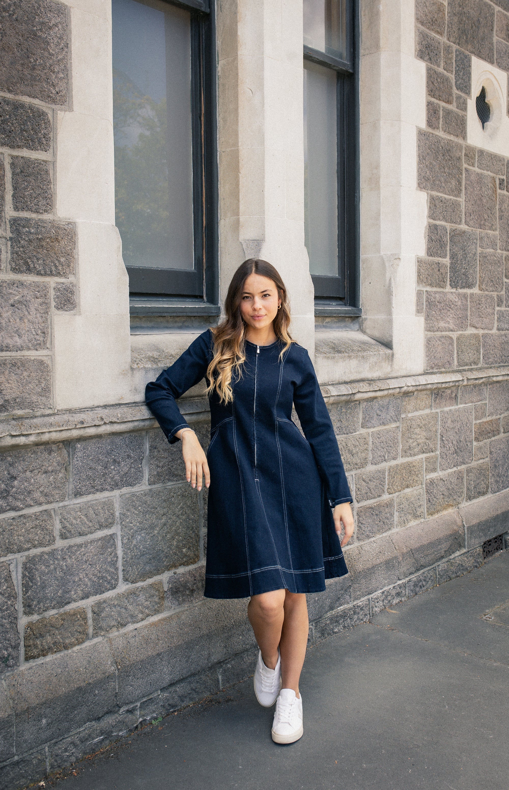 A woman with long wavy hair wearing a navy blue dress and white sneakers leans against a stone building with large windows, looking at the camera.