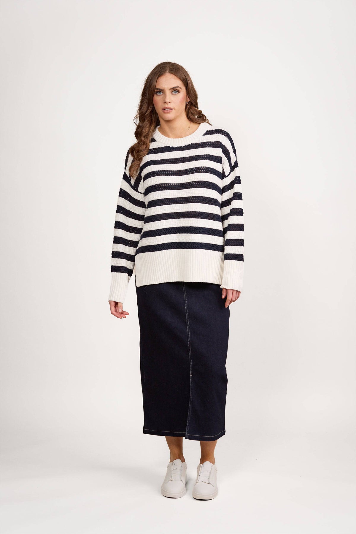 A woman with long brown hair and a neutral expression models the Vassalli Relaxed Jumper - Off White/Ink against a white background, paired with a dark denim midi skirt and white sneakers.