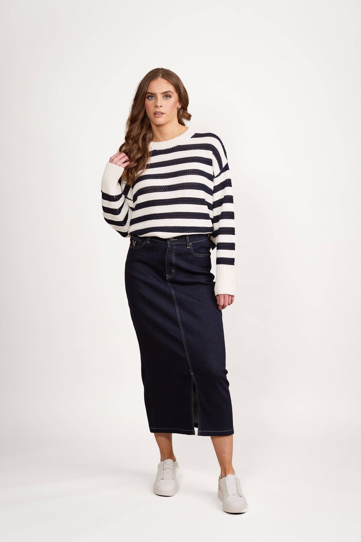 A woman with long, wavy brown hair and a neutral expression wears the Vassalli Relaxed Jumper - Off White/Ink, a dark blue denim midi skirt with a front slit, and white sneakers against a white background.