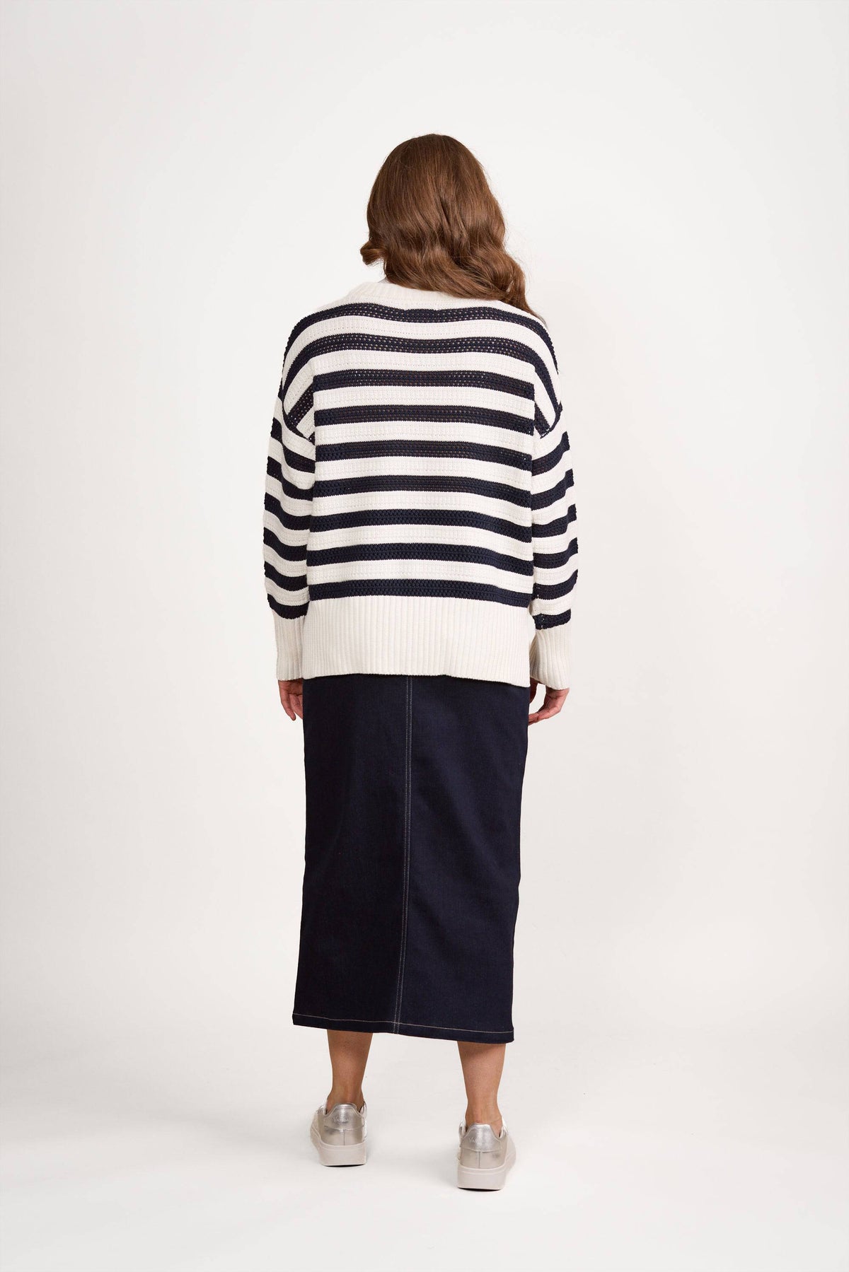 A person with wavy brown hair, wearing the Vassalli Relaxed Jumper in Off White/Ink, a dark blue midi skirt, and light-colored shoes, stands facing away against a plain white background.
