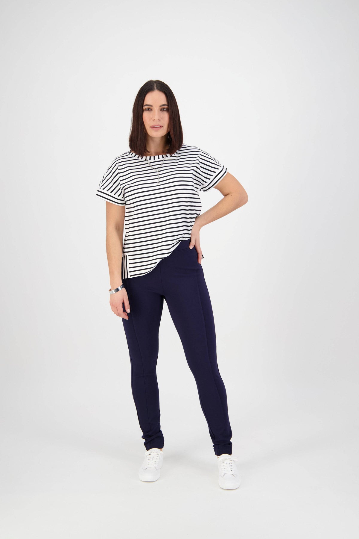 A woman with shoulder-length dark hair poses against a white background wearing the Vassalli Toll Pant in navy, a black and white striped short-sleeve top, and white sneakers, with one hand confidently on her hip.